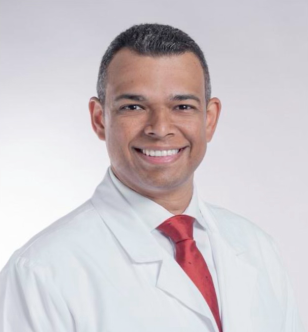 A smiling man in a white medical coat and red tie against a plain background.