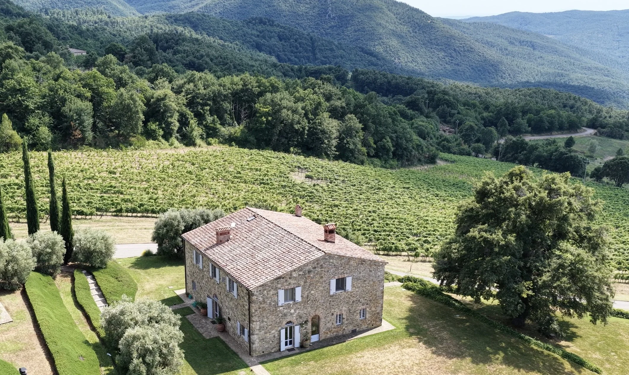 Aerial view of the stone farmhouse and terracotta roof at Castello di Fosini estate, set amidst Tuscan vineyards, cypress trees, and forested mountains.