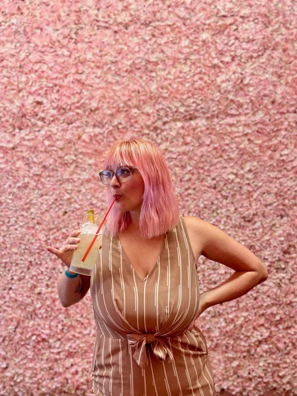 A woman with pink hair and glasses drinks a cocktail through a red straw, standing against a wall of pink flowers.