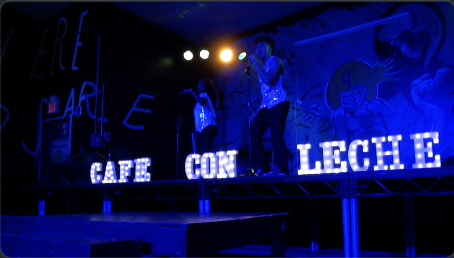 Two performers singing on a stage with blue lighting and large illuminated signs that read 'CAPE CON LECHE' and a mural in the background.