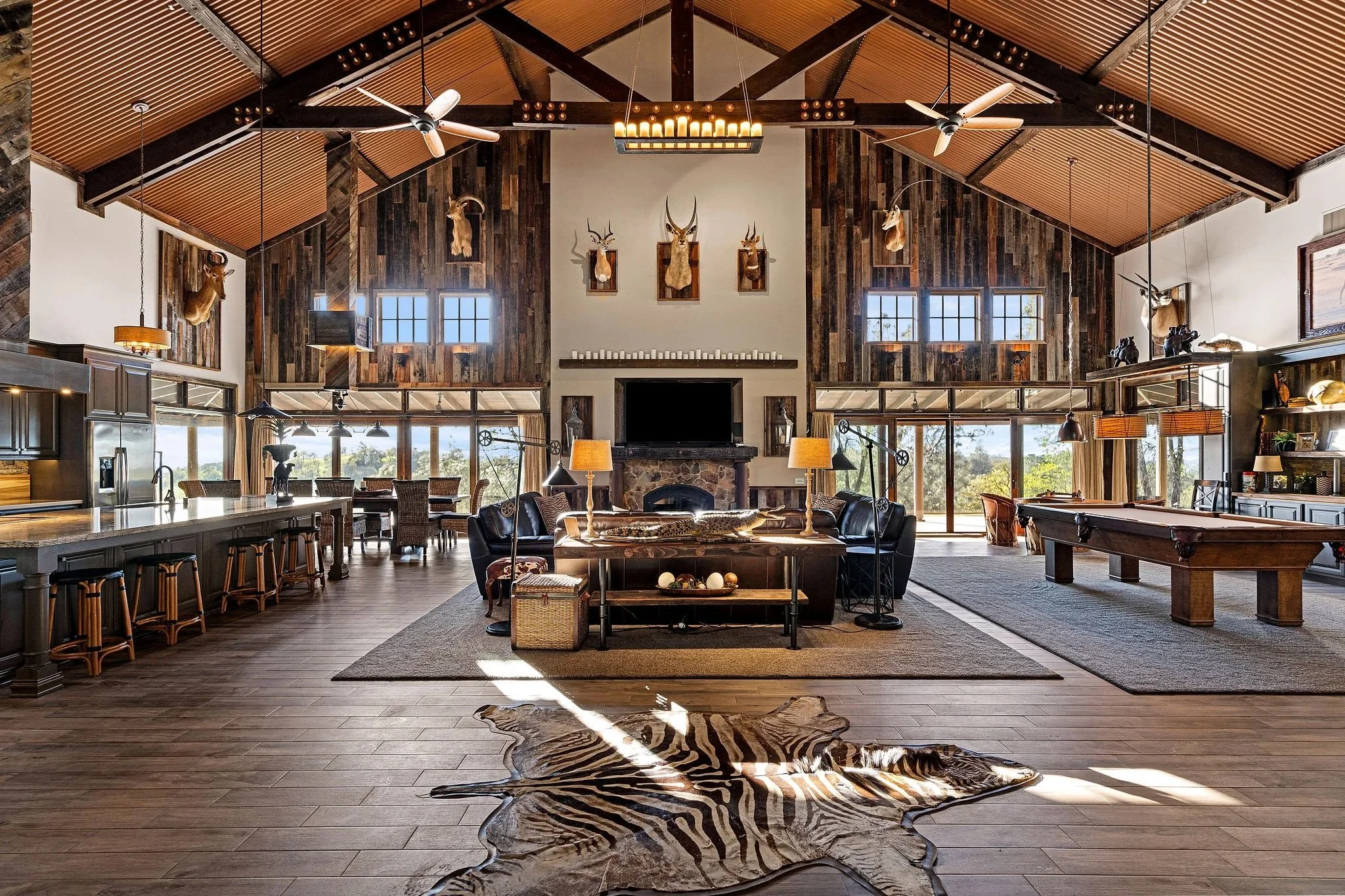Interior of a rustic lodge-style living room with high wooden ceiling, fans, and mounted animal heads. Large windows, a stone fireplace, and various furniture including couches, chairs, a pool table, and lamps. There is a zebra-patterned animal hide on the wooden floor.