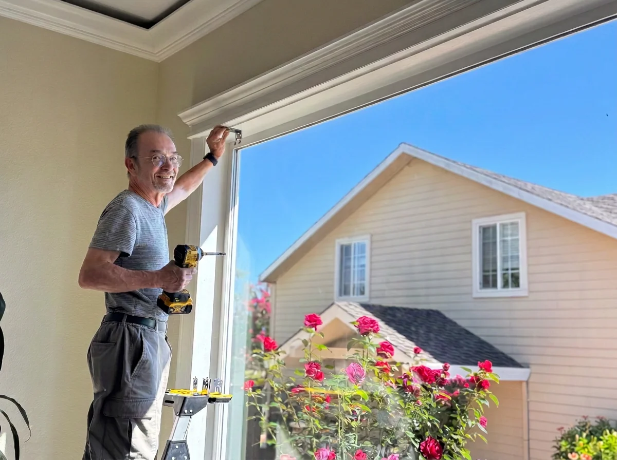 A smiling man with glasses, wearing a gray striped t-shirt and cargo shorts, is installing a window handle with a cordless drill in a room with light-colored walls. Outside the window, there is a house with beige siding, white trim, and pink flowers in the garden.
