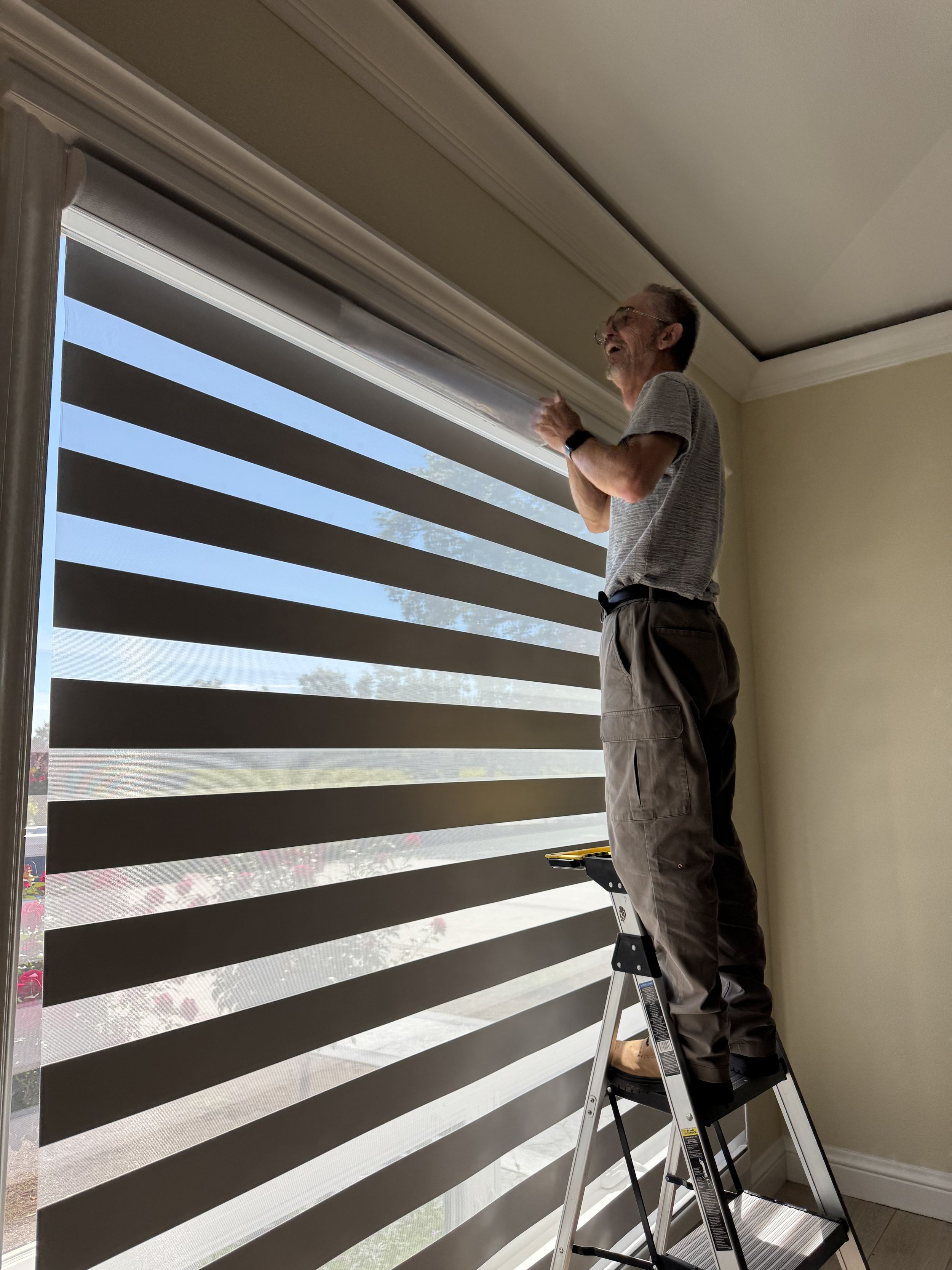 A man standing on a ladder adjusting window blinds inside a house. The blinds have horizontal stripes, partially open to let in light. The man is wearing a gray shirt, khaki pants, glasses, and a smartwatch.