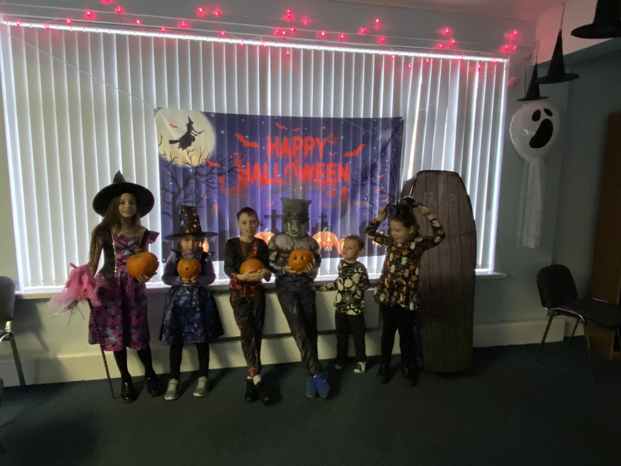 Children dressed in Halloween costumes holding pumpkins and standing in front of a Halloween themed backdrop with a spooky night scene, a witch on a broom, a full moon, and the words 'Happy Halloween' in red letters. Decorations include hanging ghost