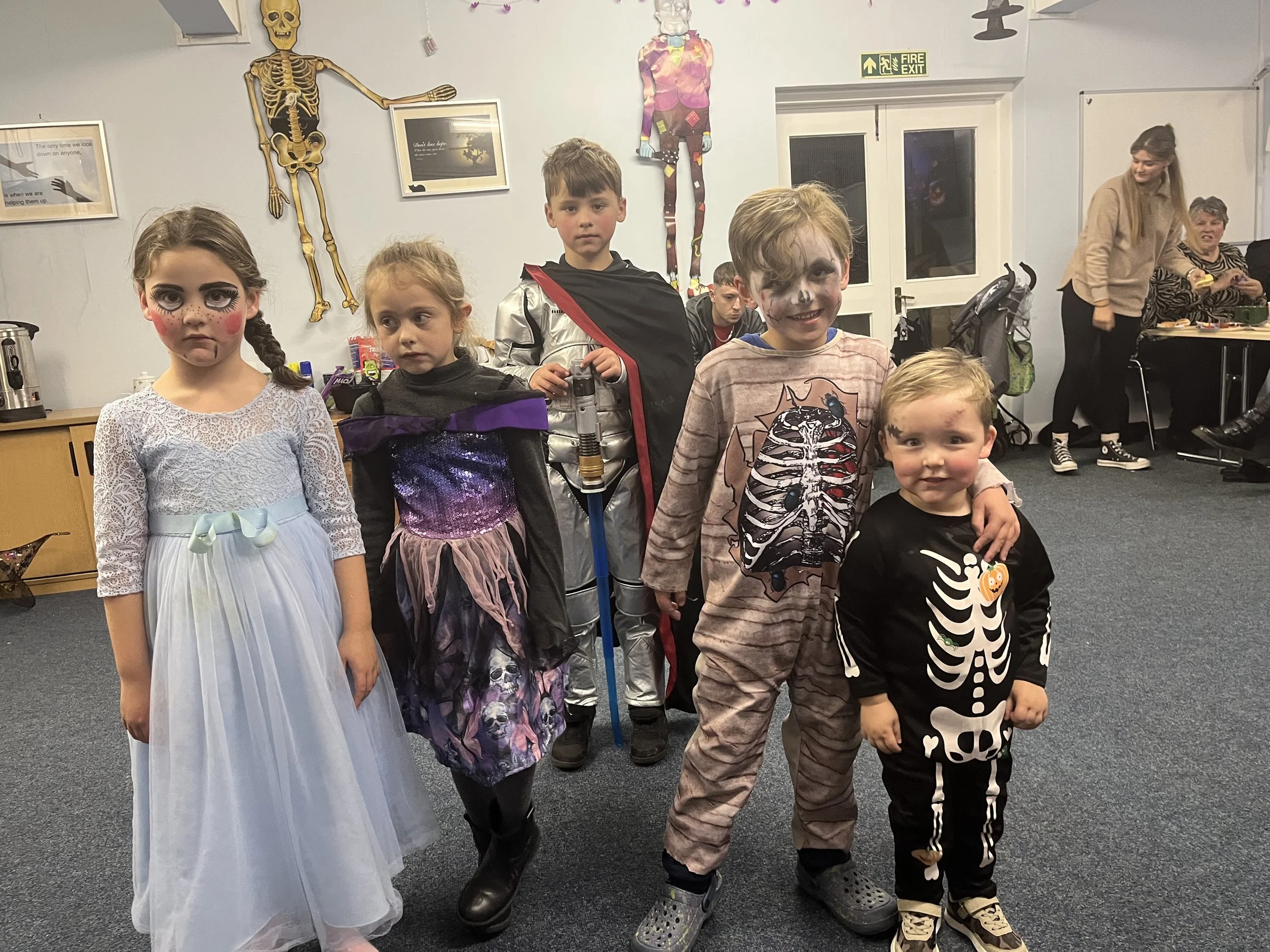Children in Halloween costumes standing indoors with decorations, including a skeleton and colorful artwork on the wall.