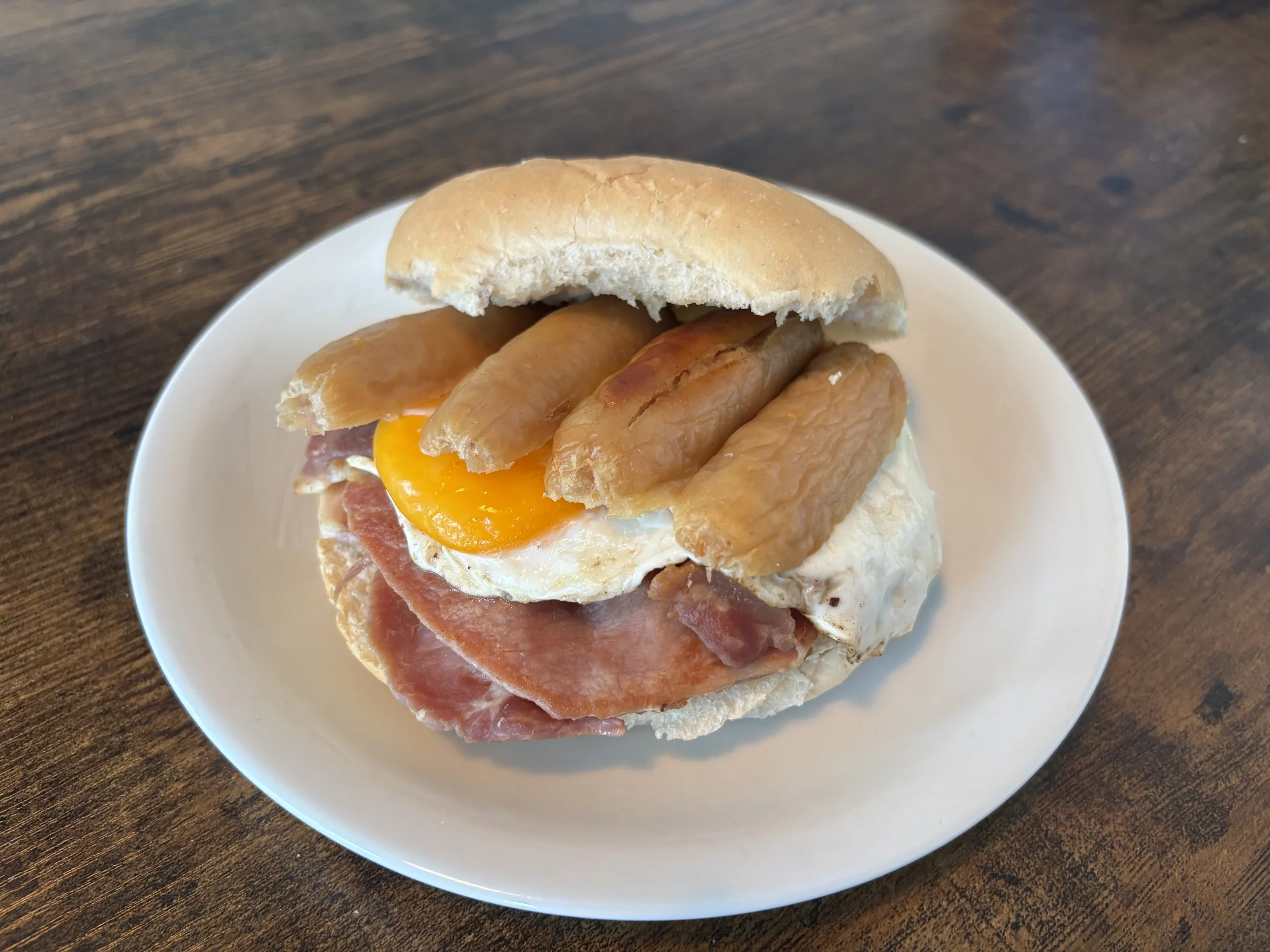 A sandwich with a fried egg, slices of ham, grilled chicken, and a sandwich bun on a white plate on a wooden table.