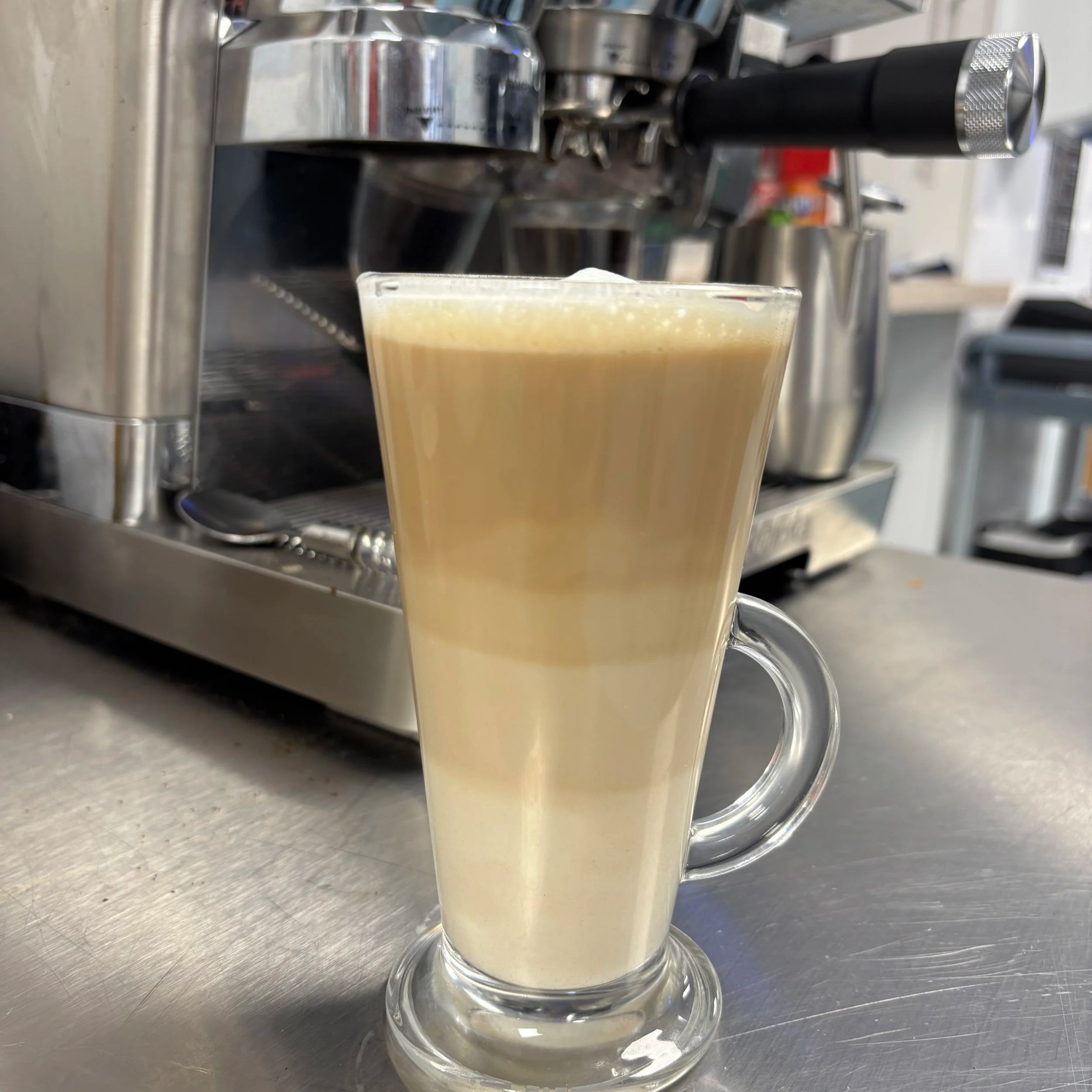 A clear glass mug with a handle on a metallic surface, filled with a frothy, light-colored coffee drink.
