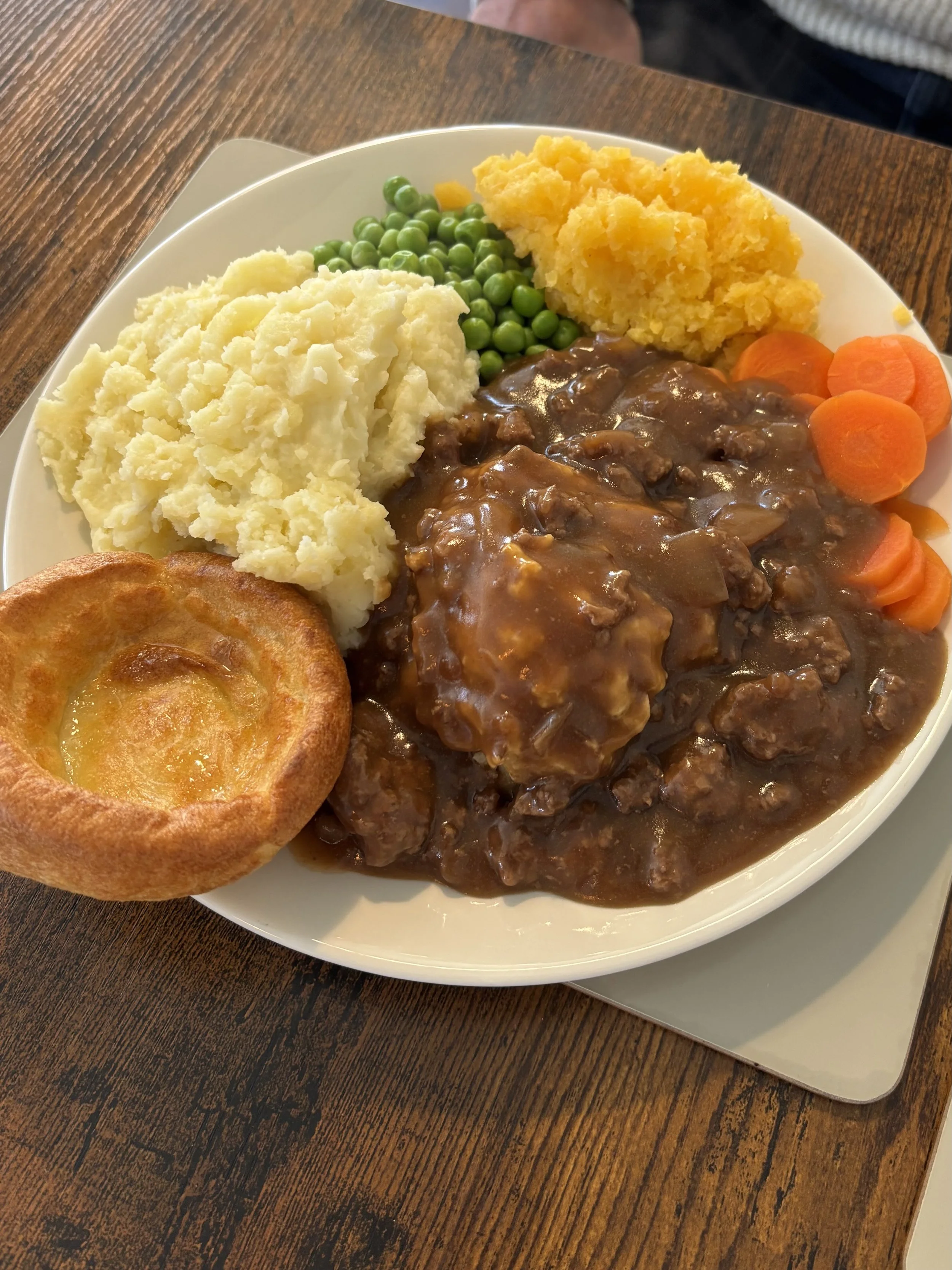 Plate of food with mashed potatoes, green peas, a serving of meat with gravy, cooked carrots, a baked bread roll, and a serving of yellow mashed foods, on a wooden table.