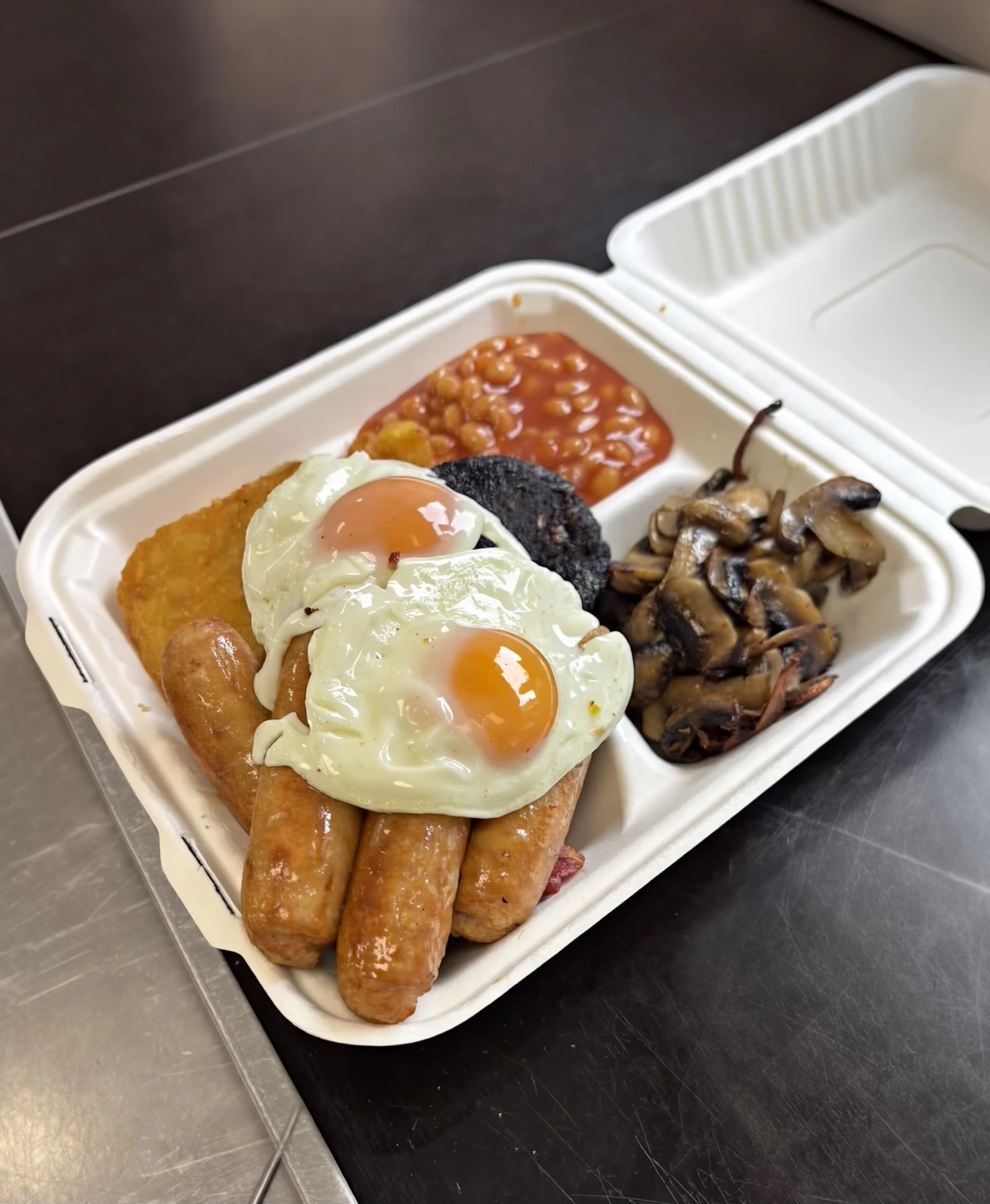 A styrofoam takeout container with baked beans, a breaded fish fillet topped with a fried egg, three sausage links, and sautéed mushrooms, placed on a black surface.