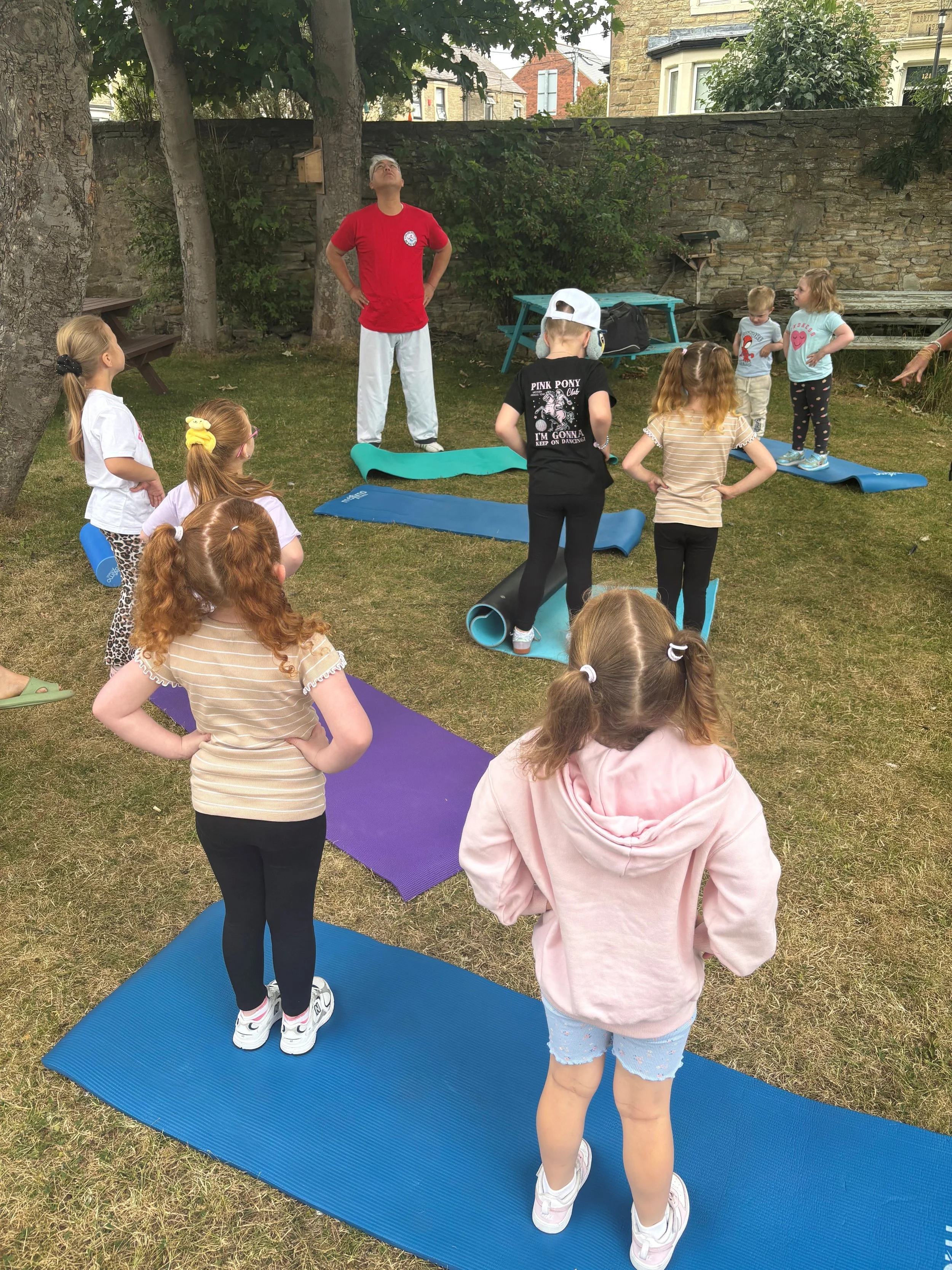A group of children participating in an outdoor yoga or exercise class on yoga mats in a garden or yard, with a male instructor standing in front of them.