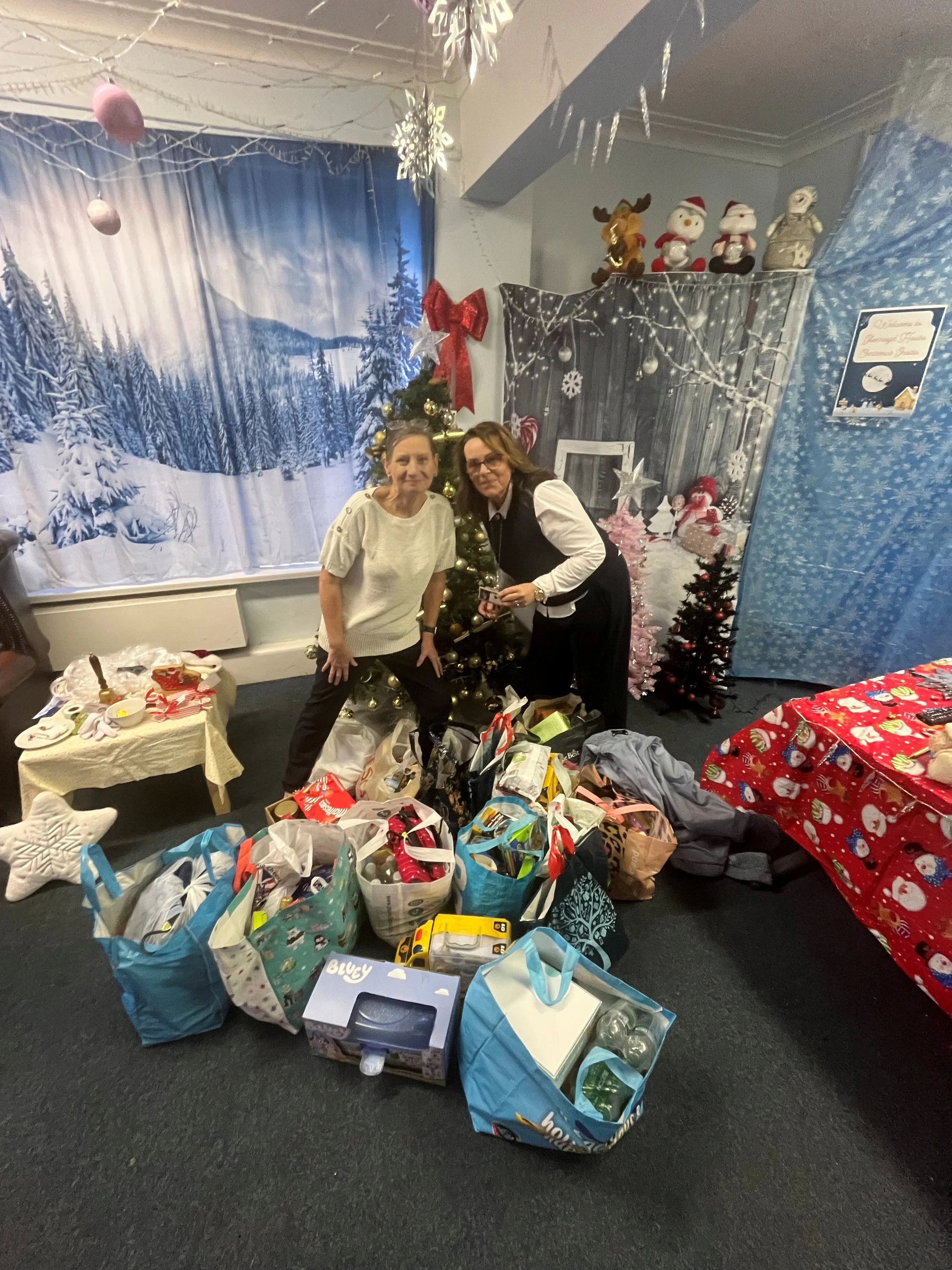 Two women standing in front of a decorated Christmas tree surrounded by wrapped gifts and shopping bags, with Christmas-themed decorations and a winter scenic backdrop in the background.