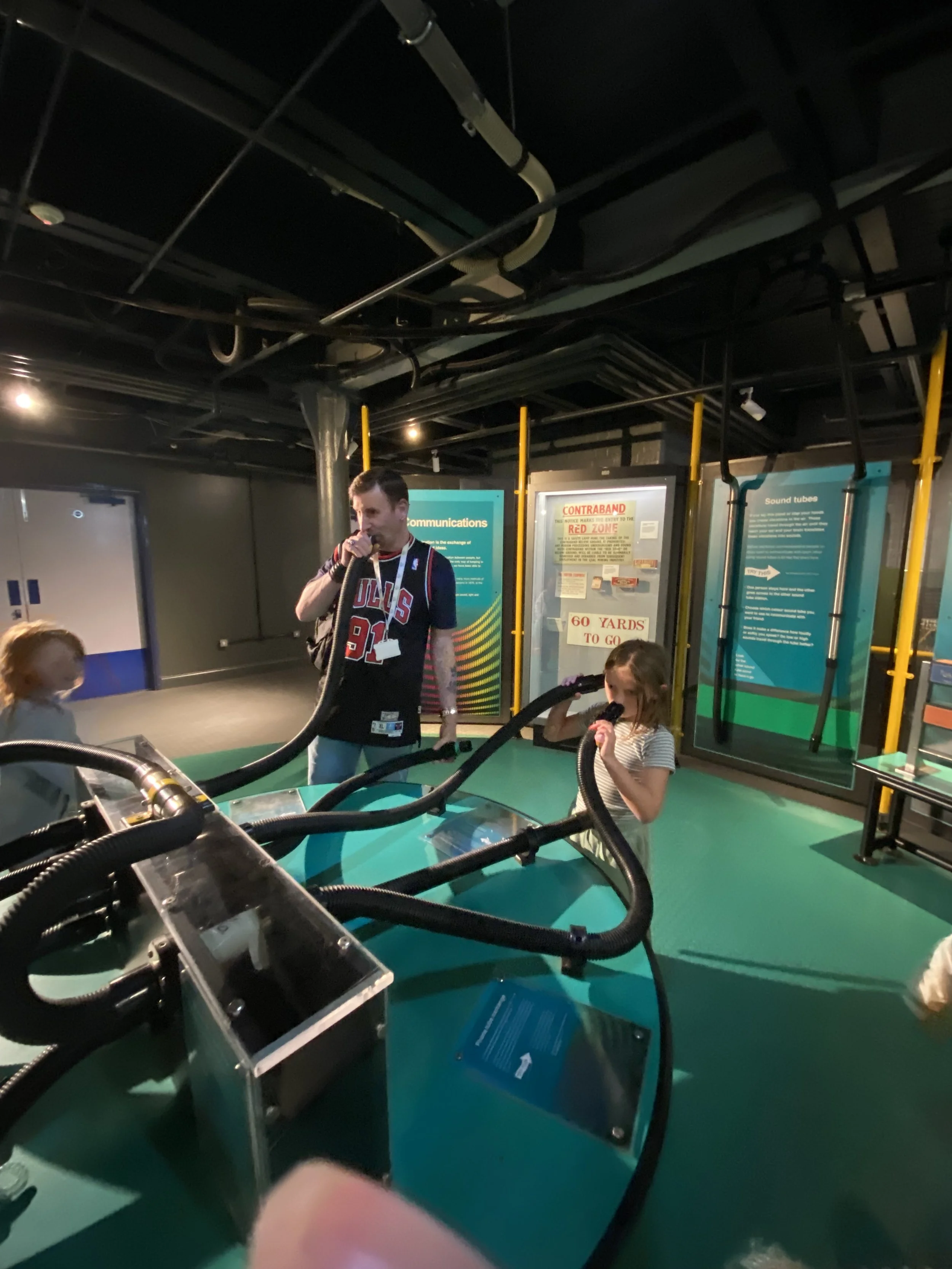 A man in a sports jersey and children in a room with informational displays, tubes, and equipment related to science or technology exhibit.