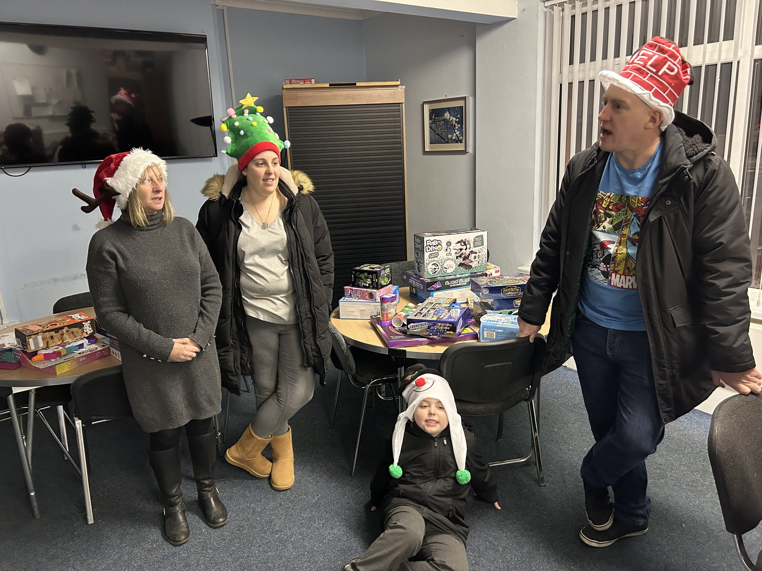 Group of five people in a room, some wearing Christmas hats, with boxes of toys on a table, and a young child sitting on the floor in a festive hat and costume.