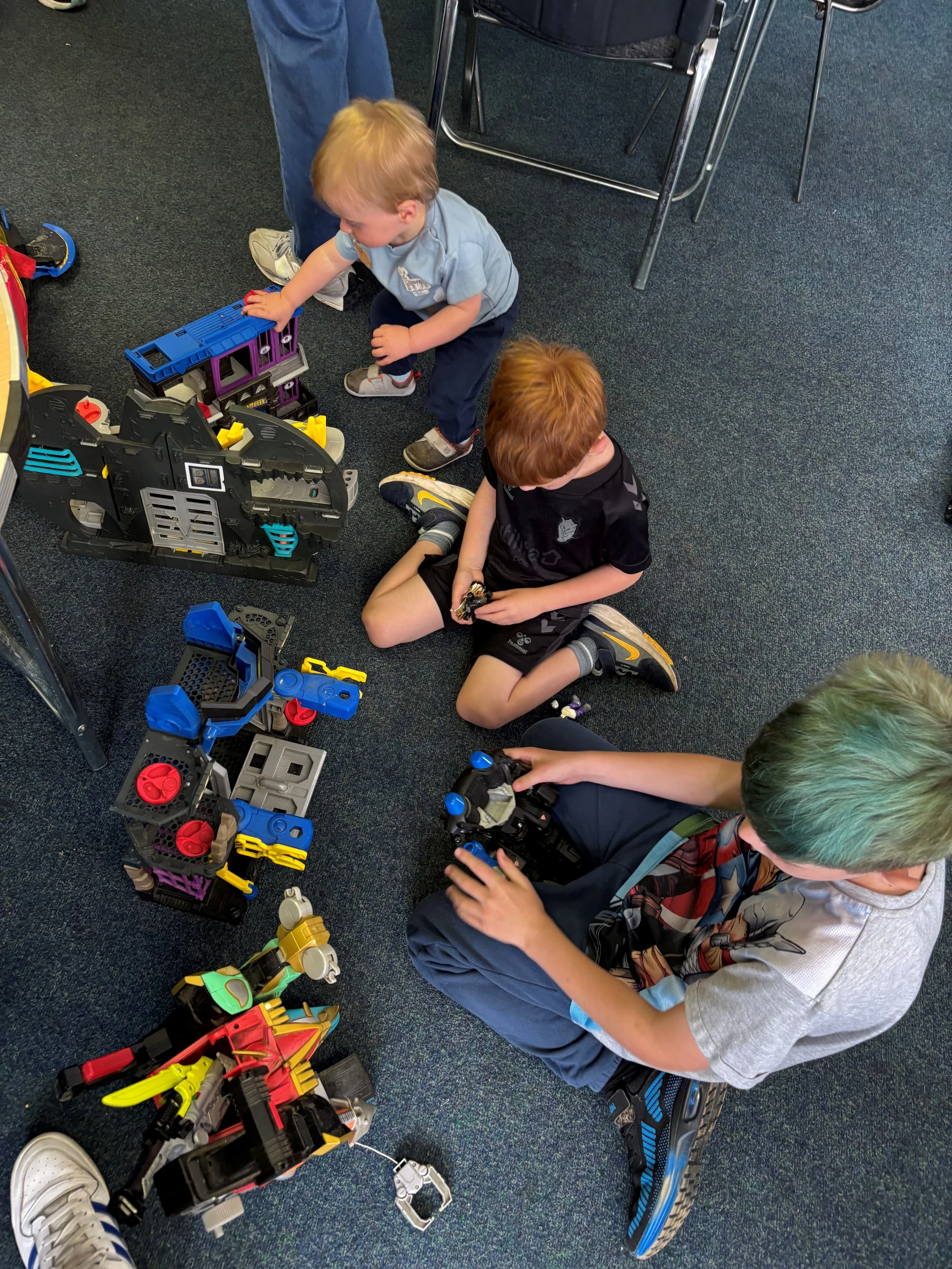 Children playing on the floor with colorful building blocks and toys, with chairs and a person standing nearby.