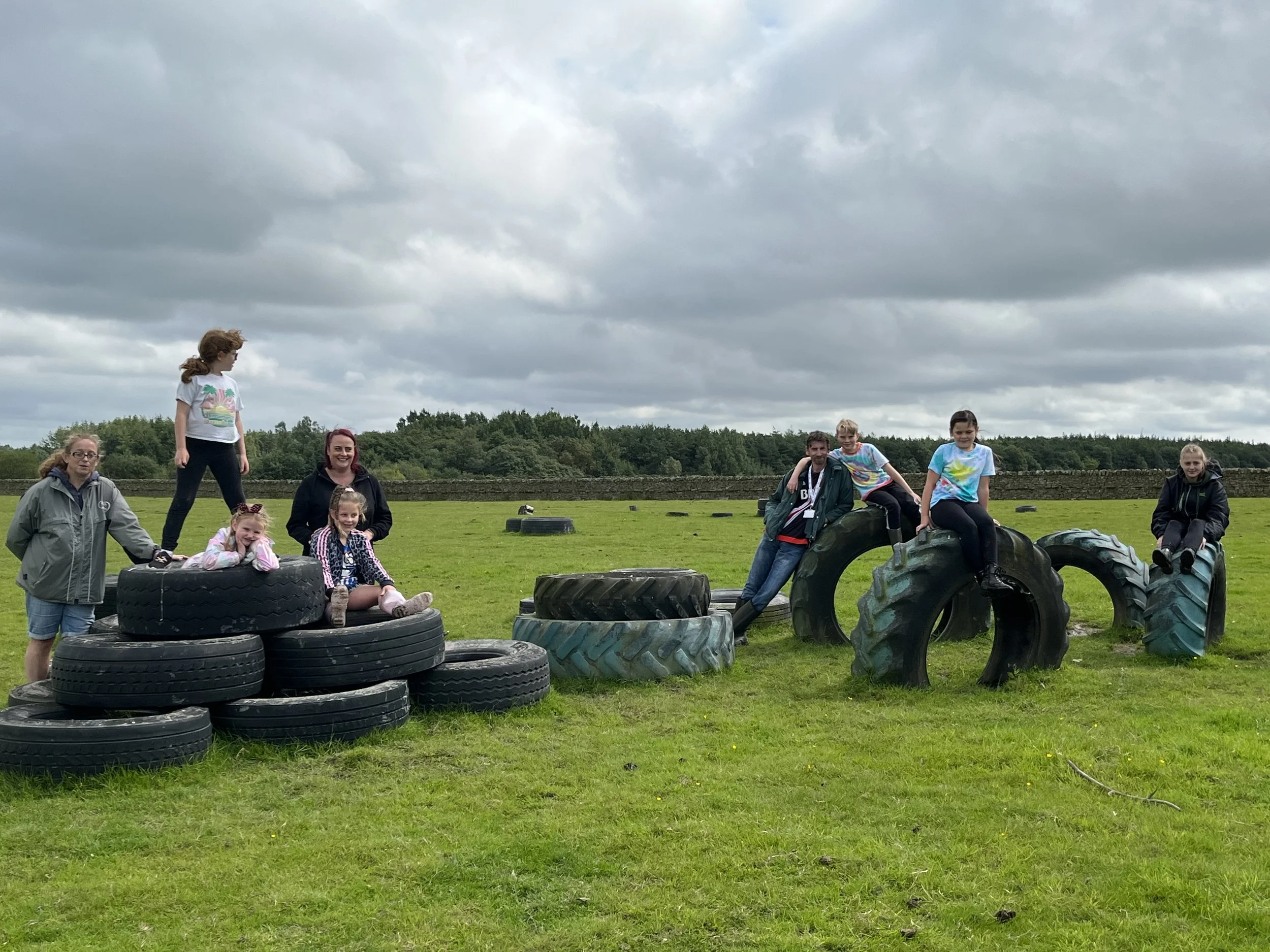 People, including children and adults, are gathered outdoors in a field hit by dark clouds. They are sitting and standing around large tires and tractor tires, which seem to be used as playground equipment.