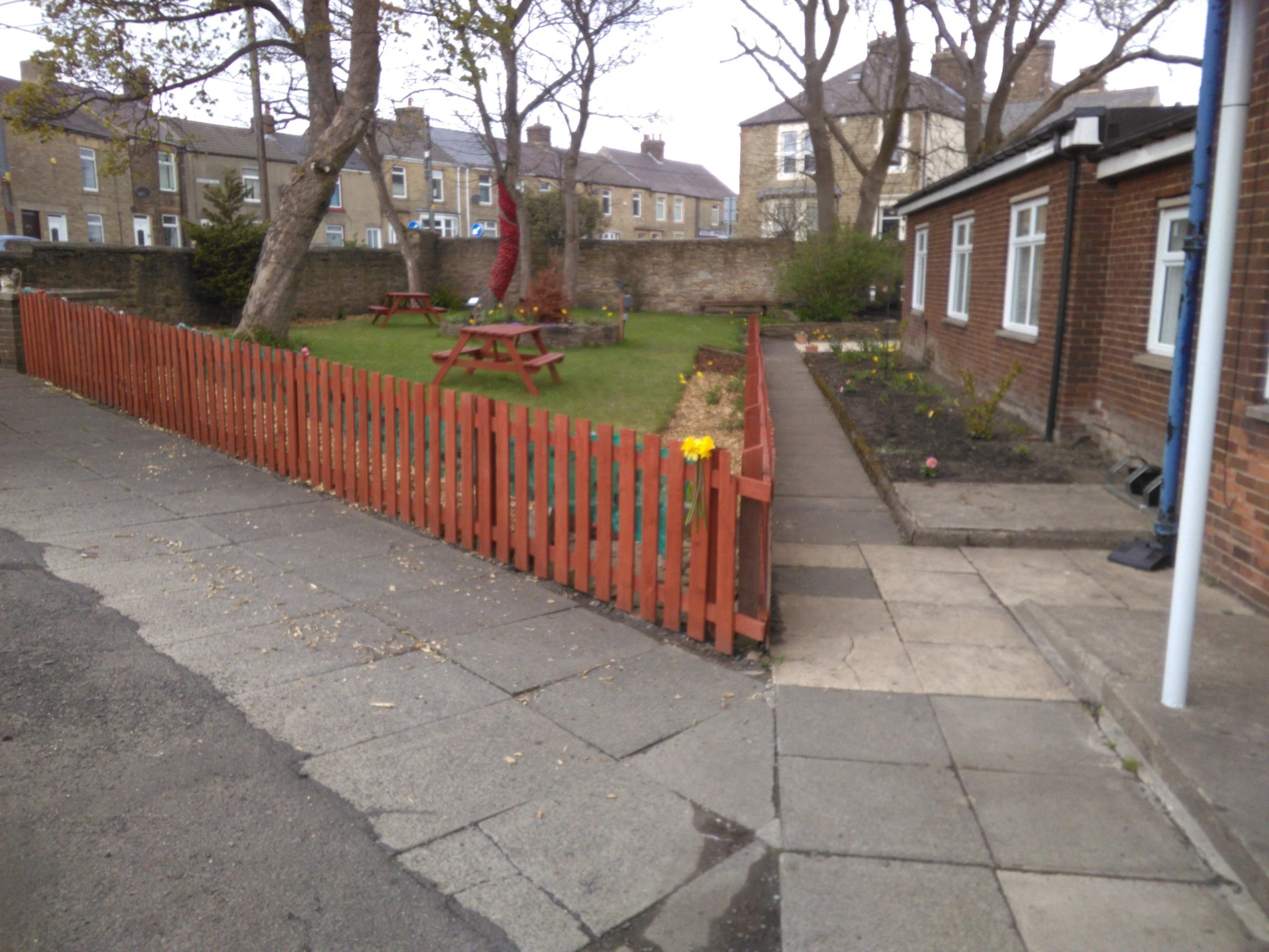 A small urban garden with a red picket fence, picnic tables, and trees behind a brick wall; surrounding buildings are residential with multiple windows.