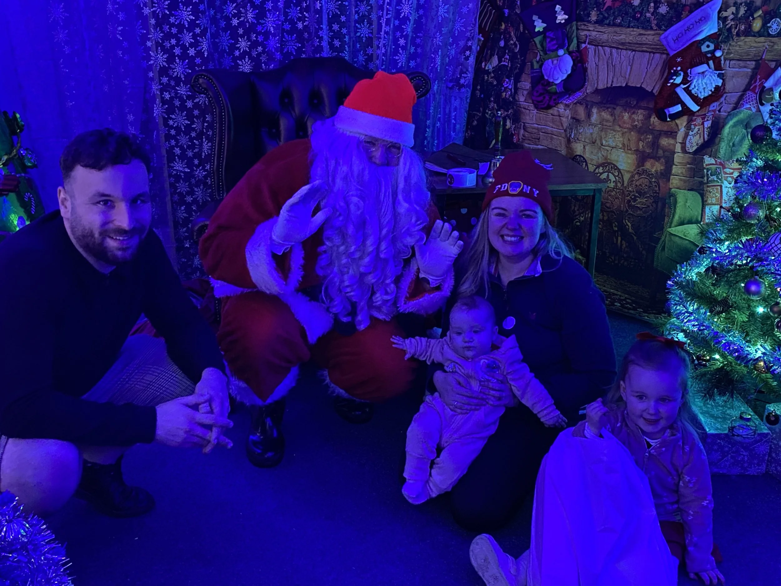 People posing with Santa Claus in a festive room decorated for Christmas, with a Christmas tree and stockings.