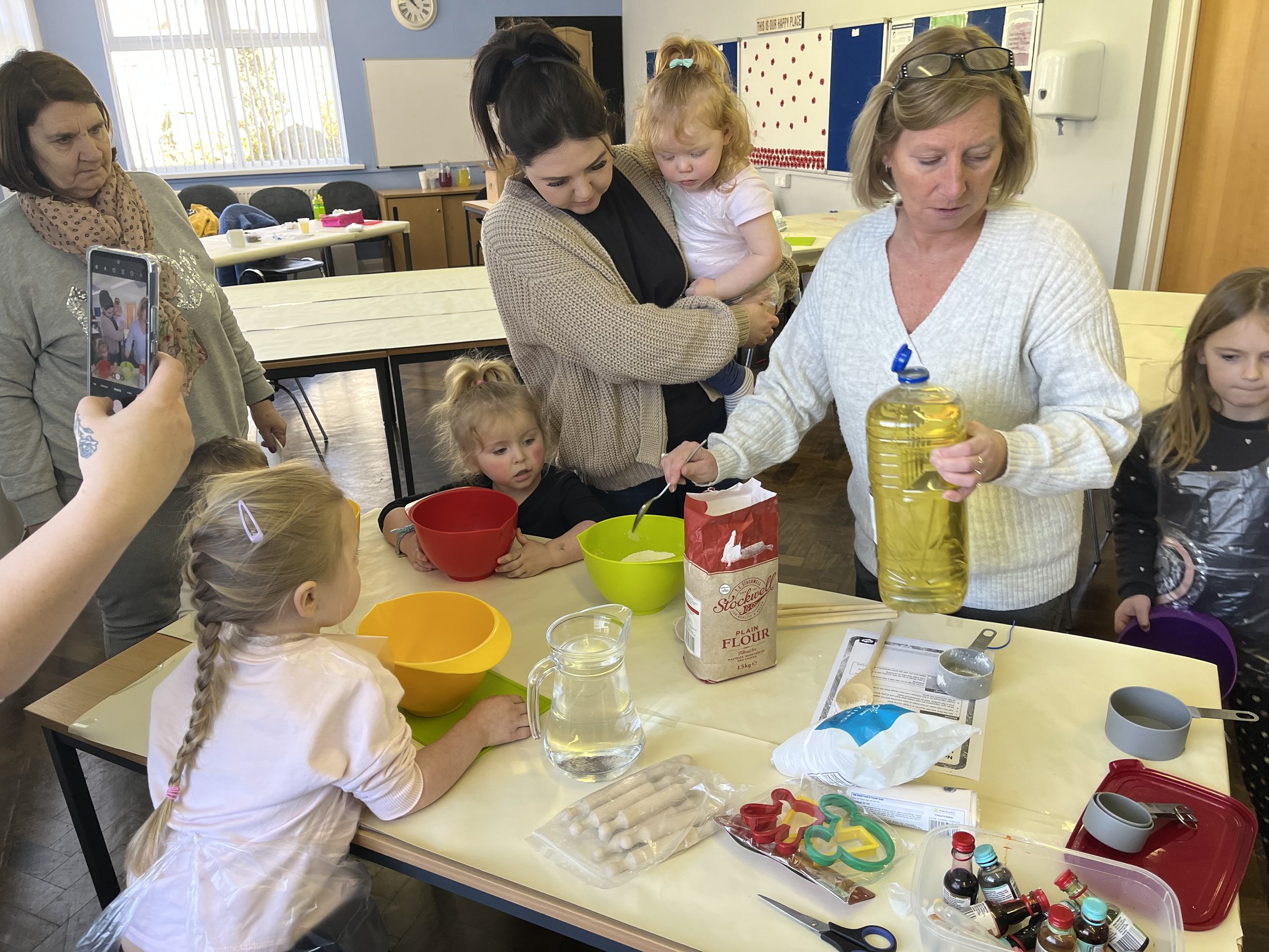 Group of children and women participating in a baking or cooking activity in a classroom, with various bowls, ingredients, and tools on the table.