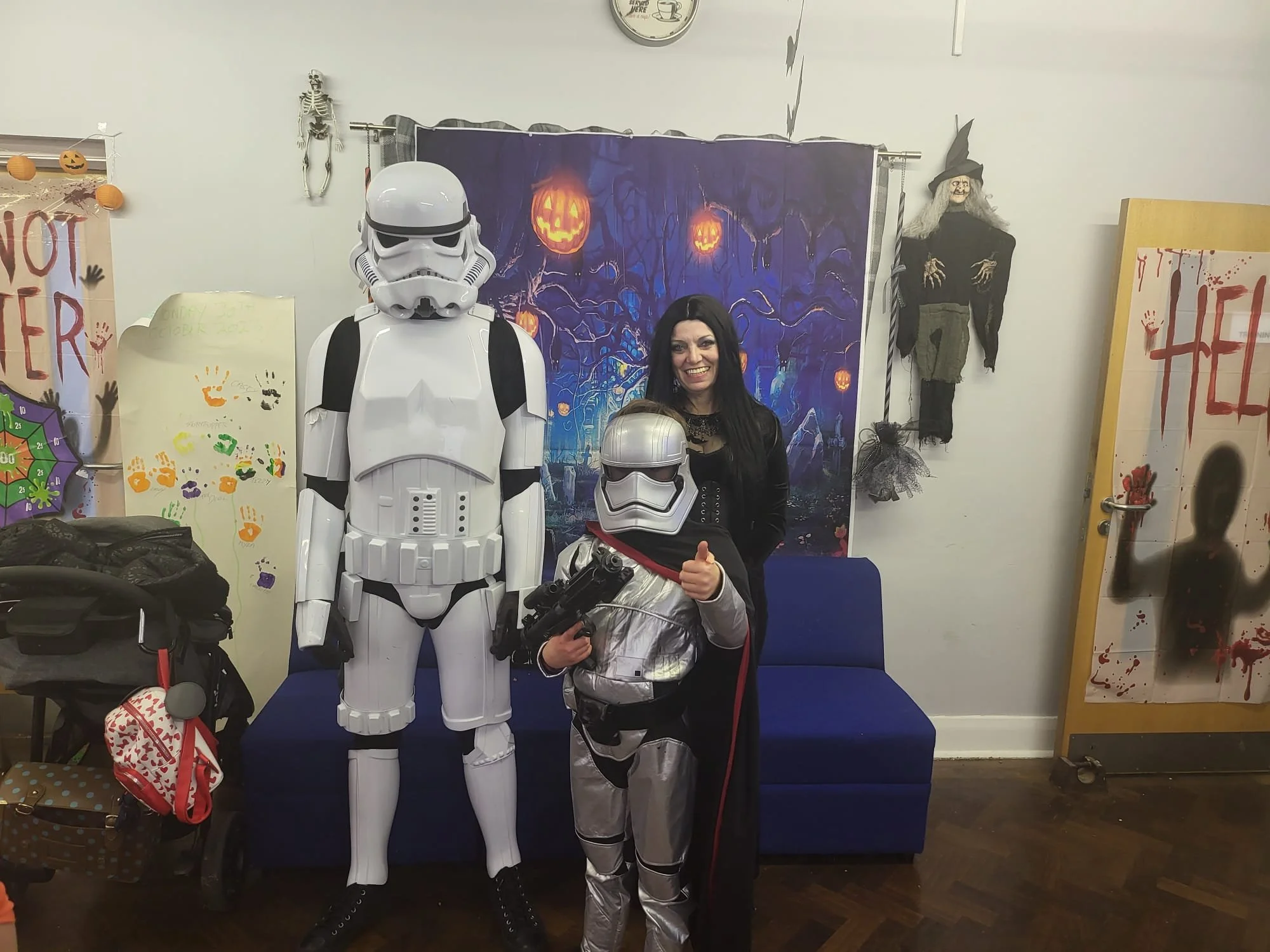 Two people dressed as characters from Star Wars, a woman with black hair and a child in a silver and black stormtrooper costume, posing with a life-sized Stormtrooper figure. The setting has Halloween decorations including a skeleton, hanging pumpkin