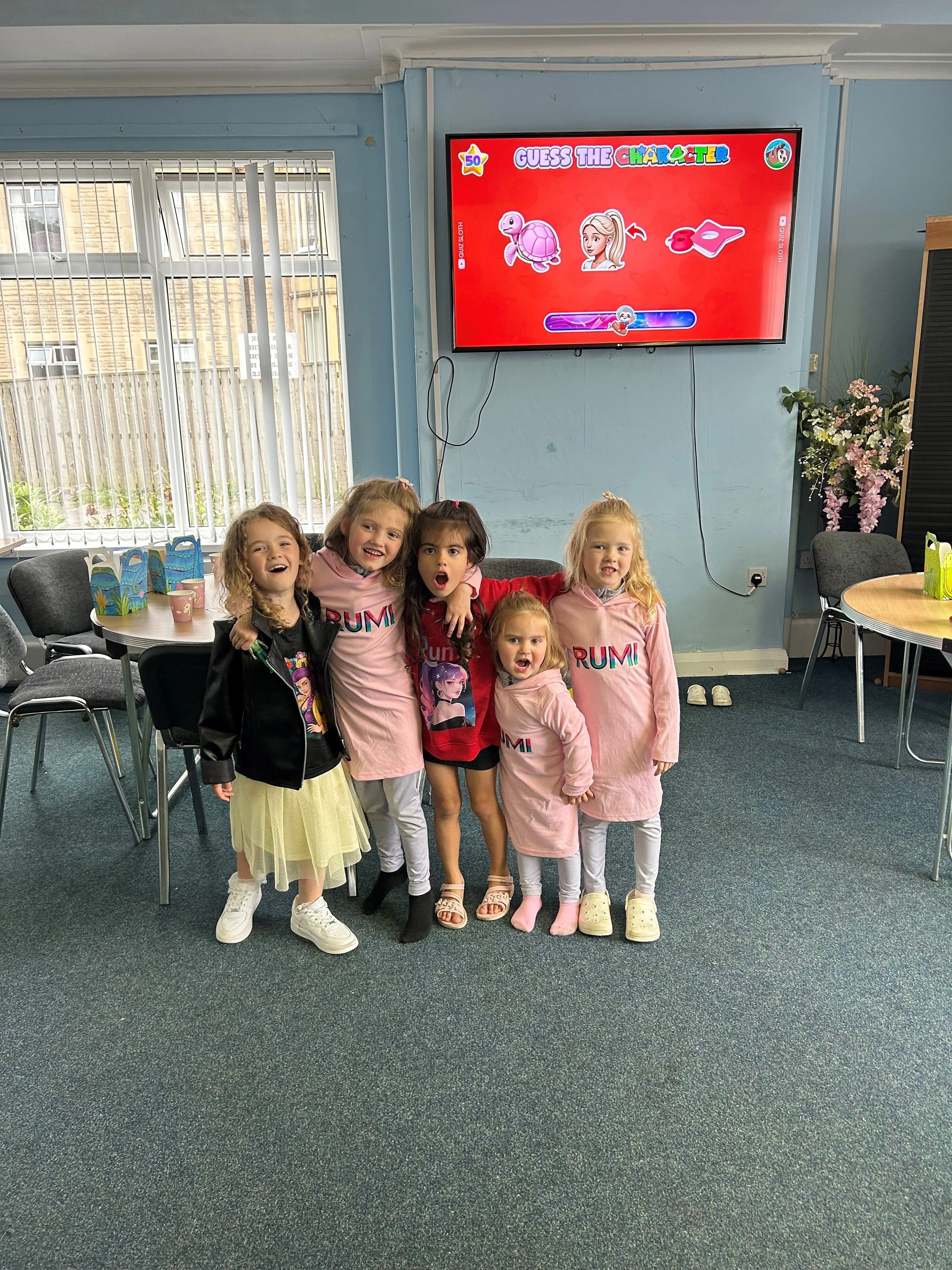 Group of six young girls standing together in a room, smiling and posing for a photo, with a TV screen displaying a game called "Guess the Character" in the background.