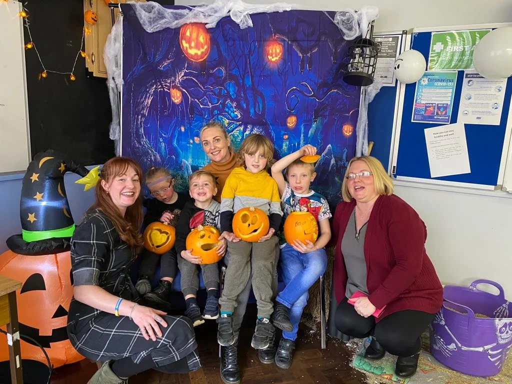Group of children and two women celebrating Halloween with pumpkins, Halloween decorations, and a spooky backdrop.