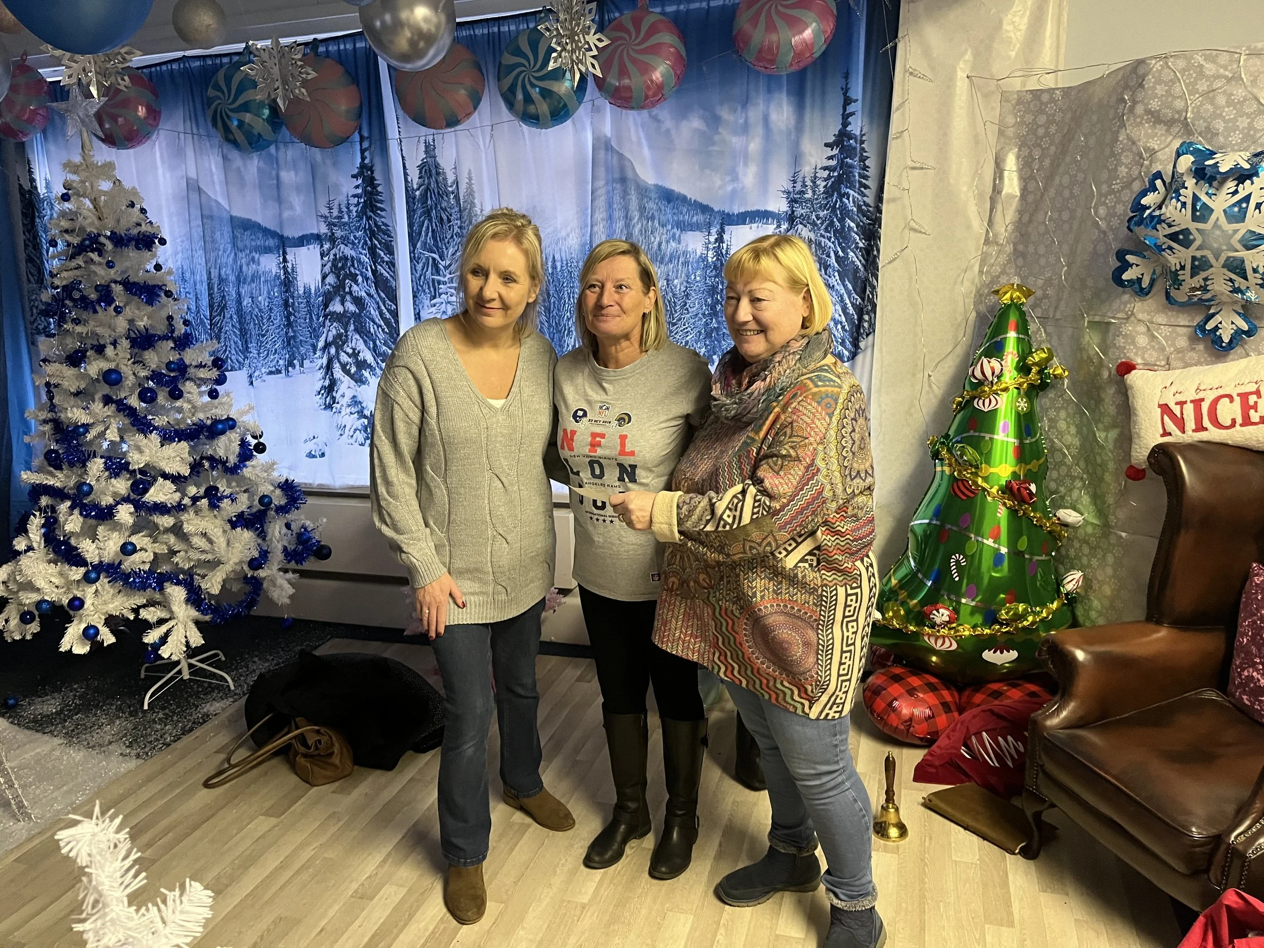 Three women standing together in front of a snowy mountain and pine forest backdrop, decorated with Christmas ornaments, trees, and holiday decor.