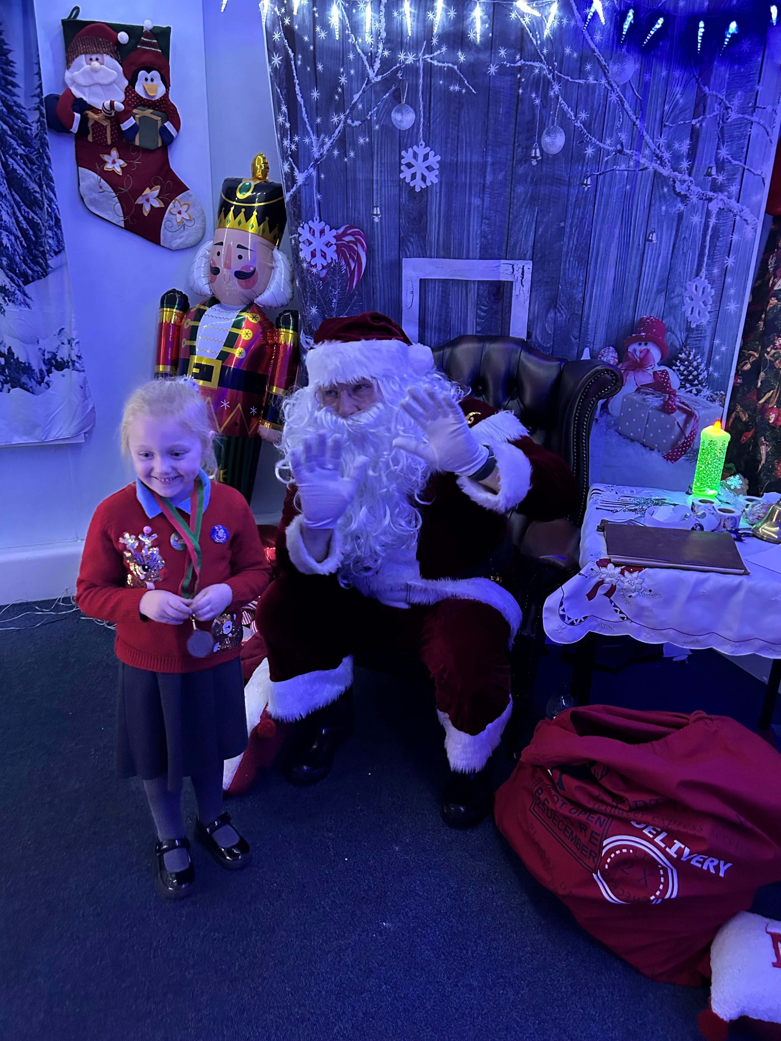 A young girl in a red Christmas sweater with medals around her neck smiling next to Santa Claus, who is dressed in a traditional red suit with white trim, sitting on a black leather chair. The background features Christmas decorations including a large inflatable nutcracker, a Christmas stocking, a snowflake, and festive winter scene artwork with snow and icicles, along with wrapped gifts and a glowing green candle on a table.