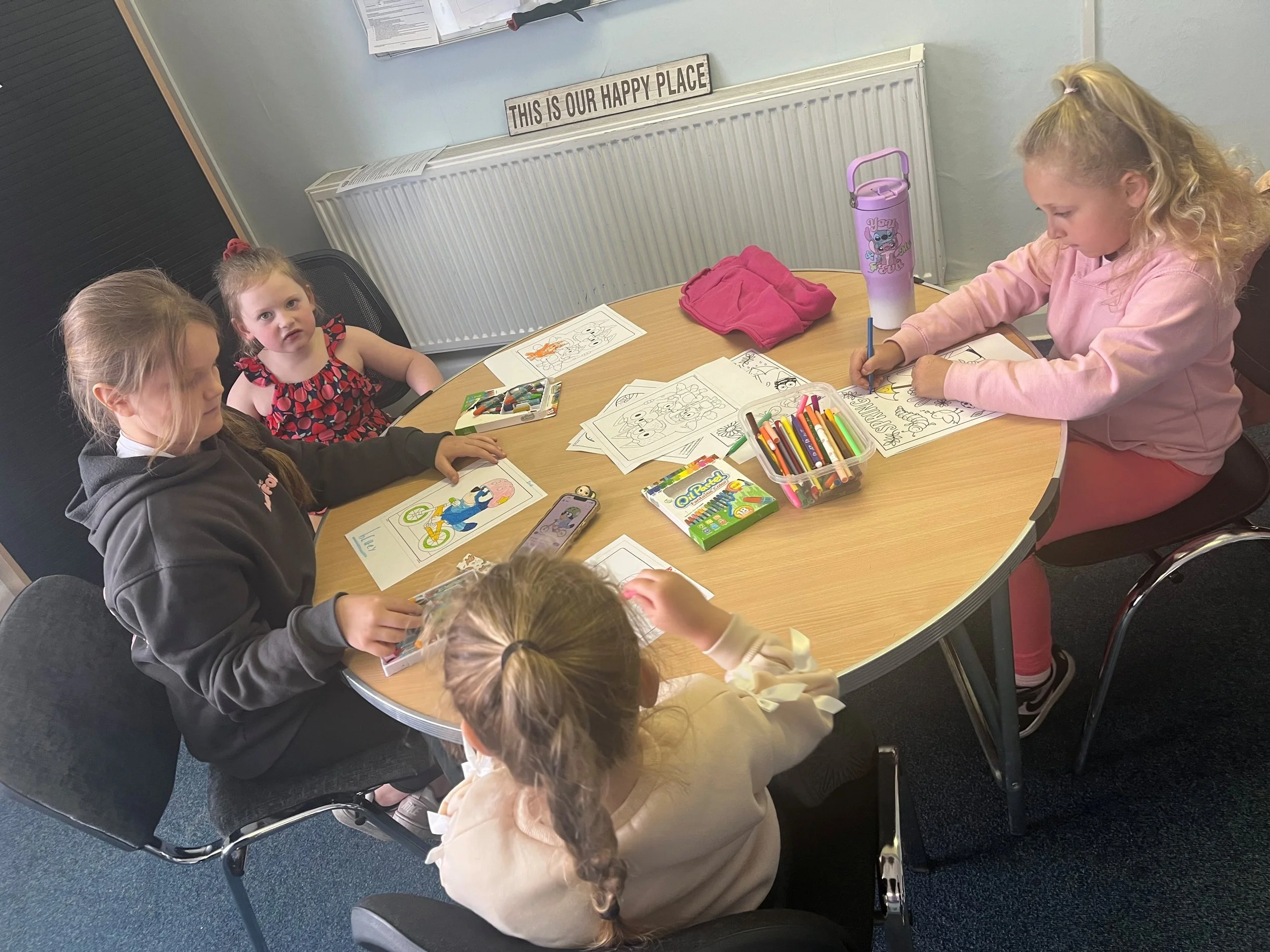 Four young girls seated at a round table engaged in coloring activities, with coloring pages and markers in front of them. One girl is wearing a pink sweatshirt, and a purple water bottle with a cartoon character is visible on the table. A decorative