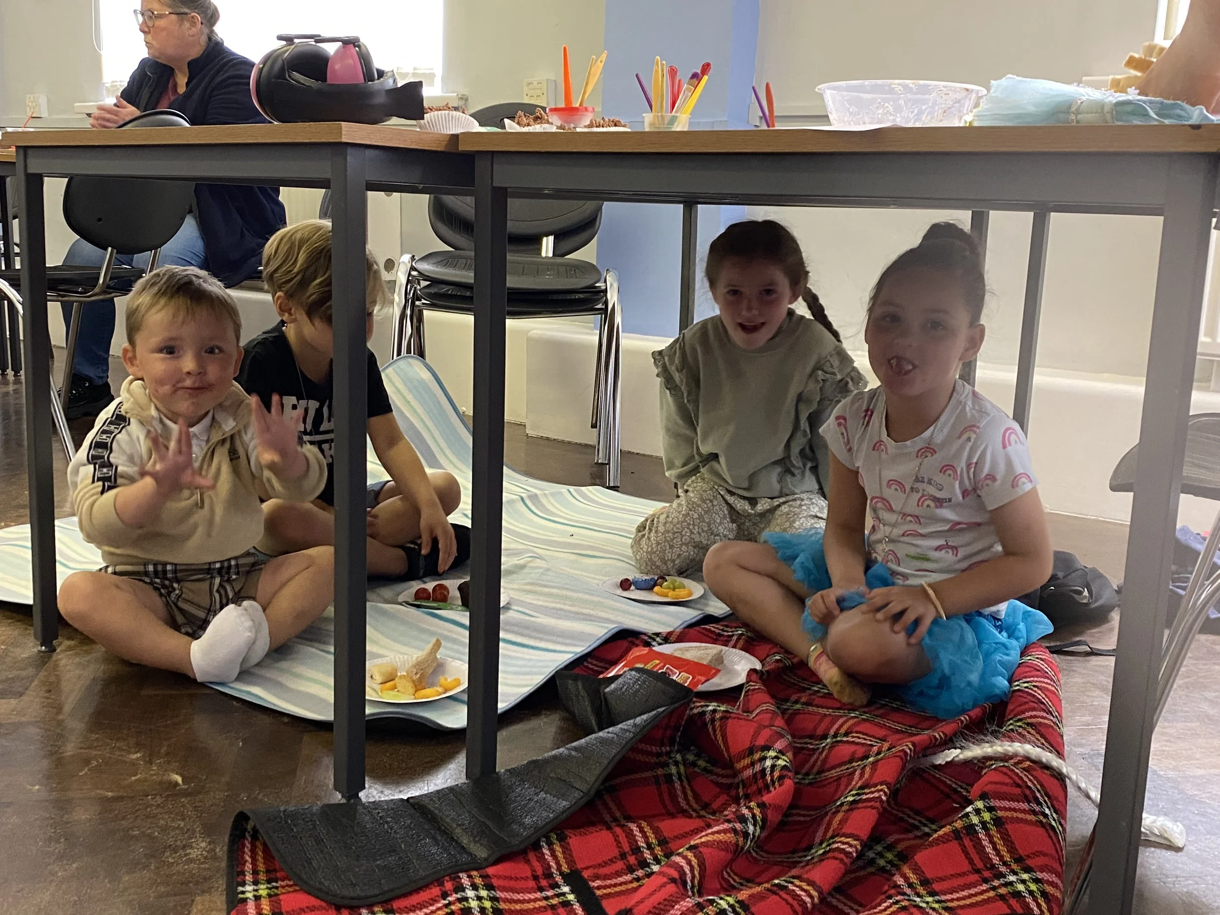 Children sitting on the floor under a table with snacks and toys, with an adult woman sitting at the table above them.