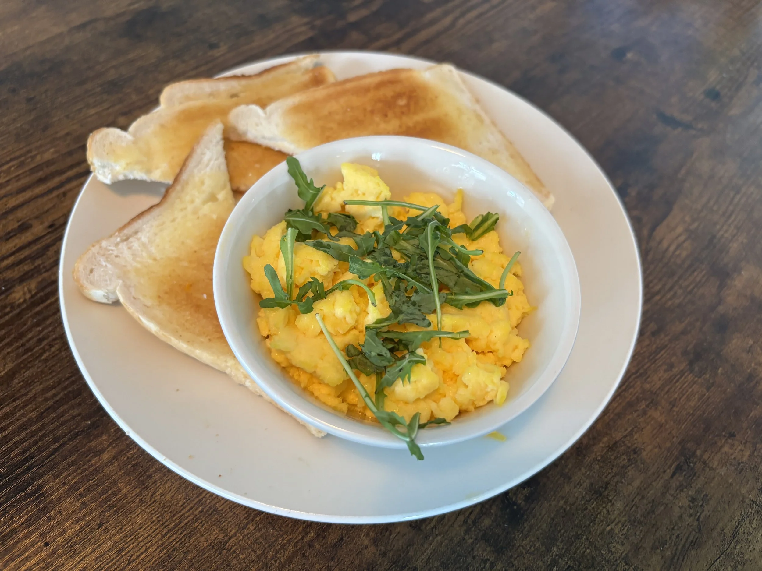 A white bowl of scrambled eggs garnished with greens on a white plate, accompanied by toasted bread slices on a dark wooden table.