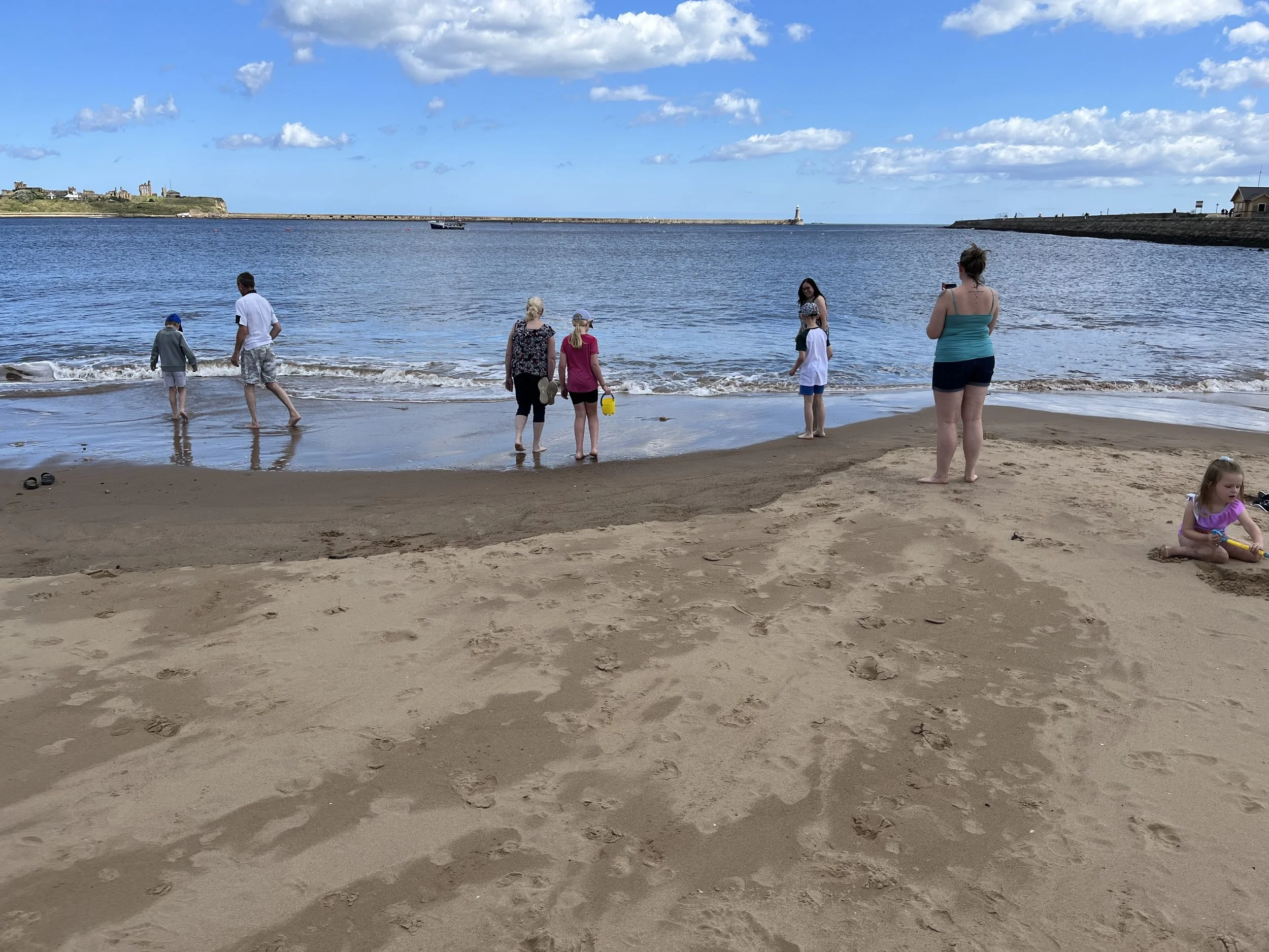 People enjoying the beach with sand, water, and a blue sky with clouds. Some individuals are wading in the water, and others are standing or sitting on the sand. In the background, there are rocks, a lighthouse, and a small structure along the shorel