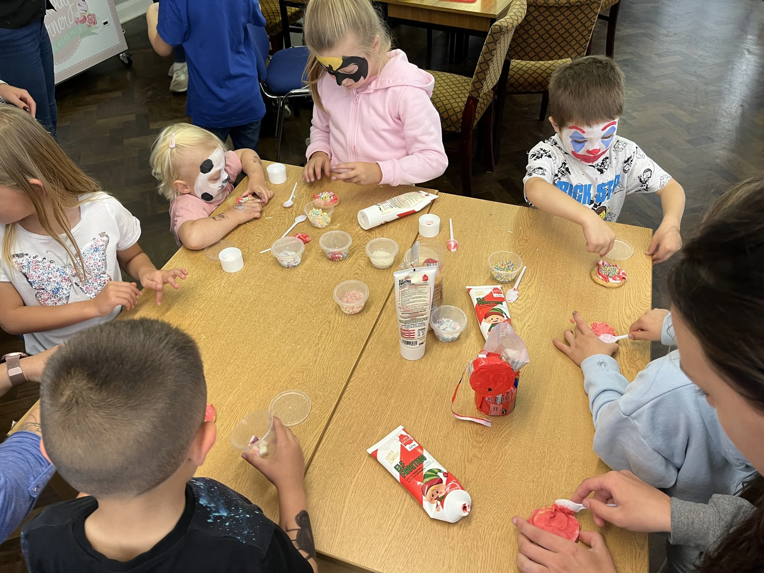 Children with face paint decorating cookies and frosting at a table with various sprinkles and decorating supplies.