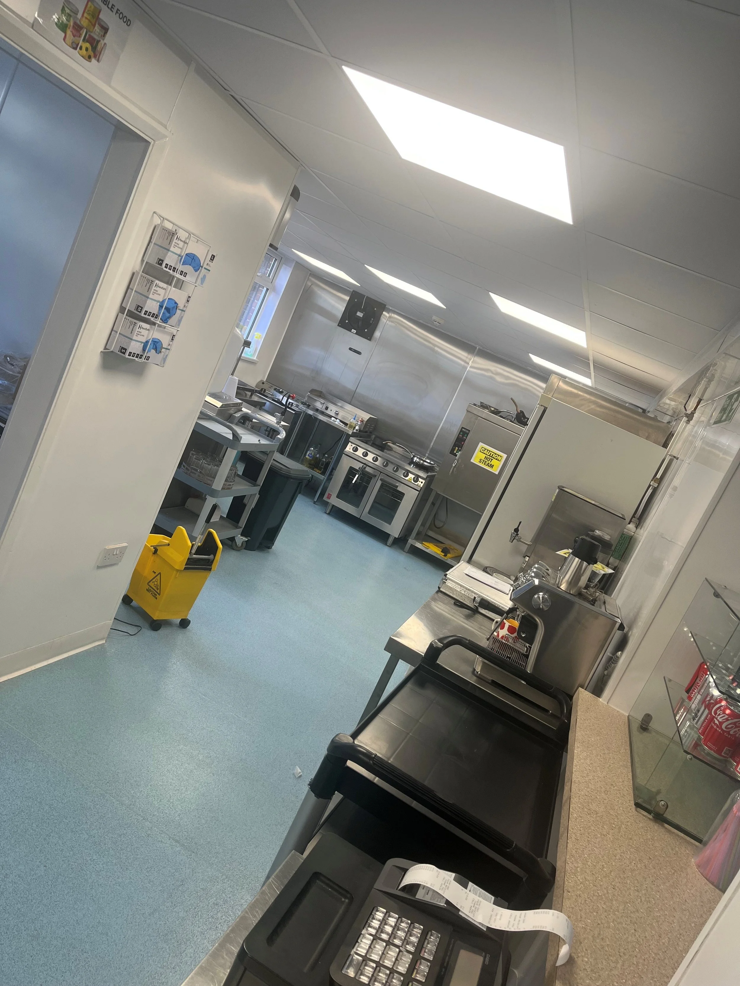 Hospital kitchen with stainless steel appliances, a cash register, and a yellow caution mop bucket on a blue floor. Ceiling lights illuminate the room.