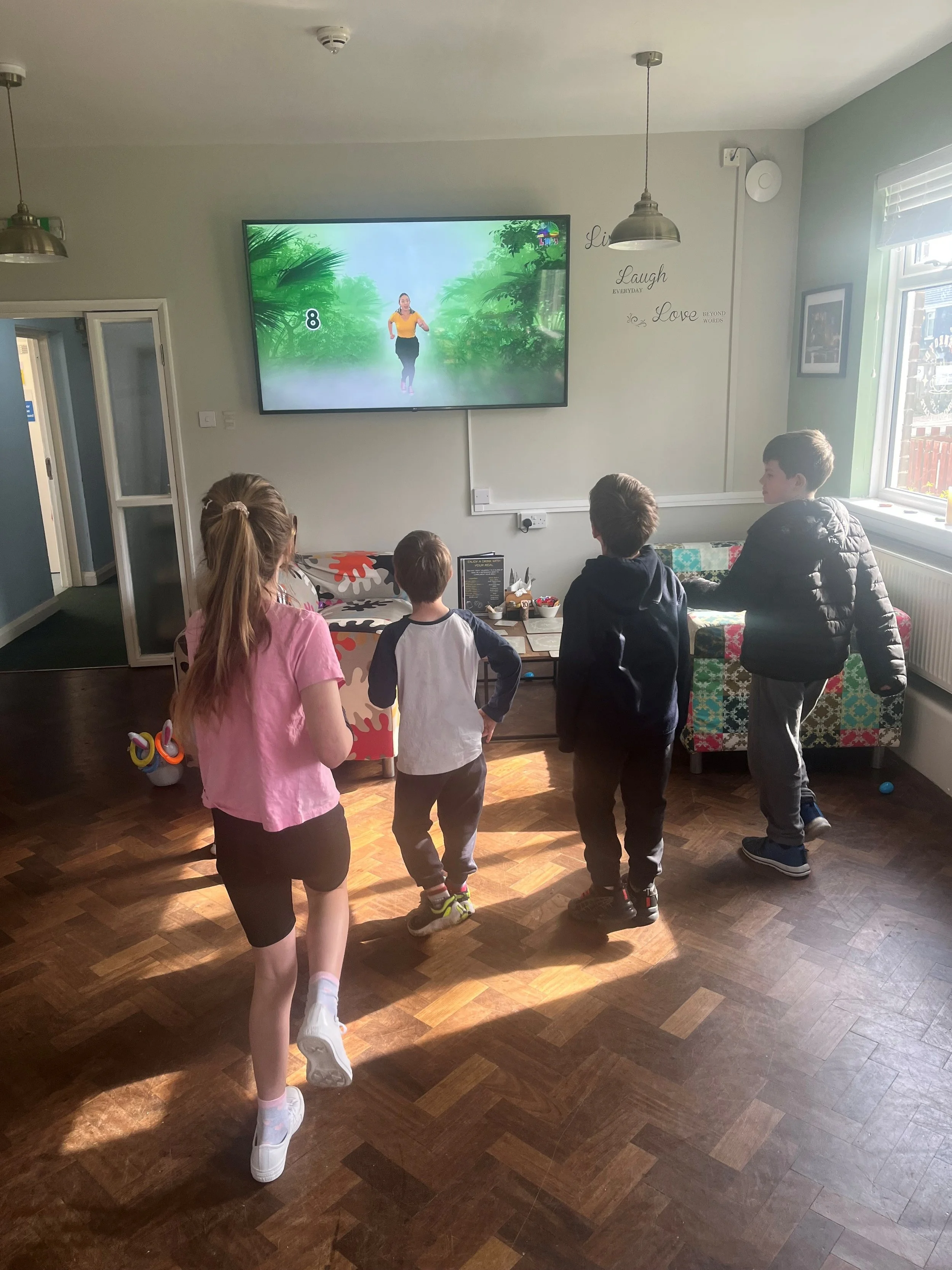 Four children, three boys and one girl, watching a woman lead an exercise class on a TV screen in a cozy living room. The woman on the TV is running in a virtual jungle scene.