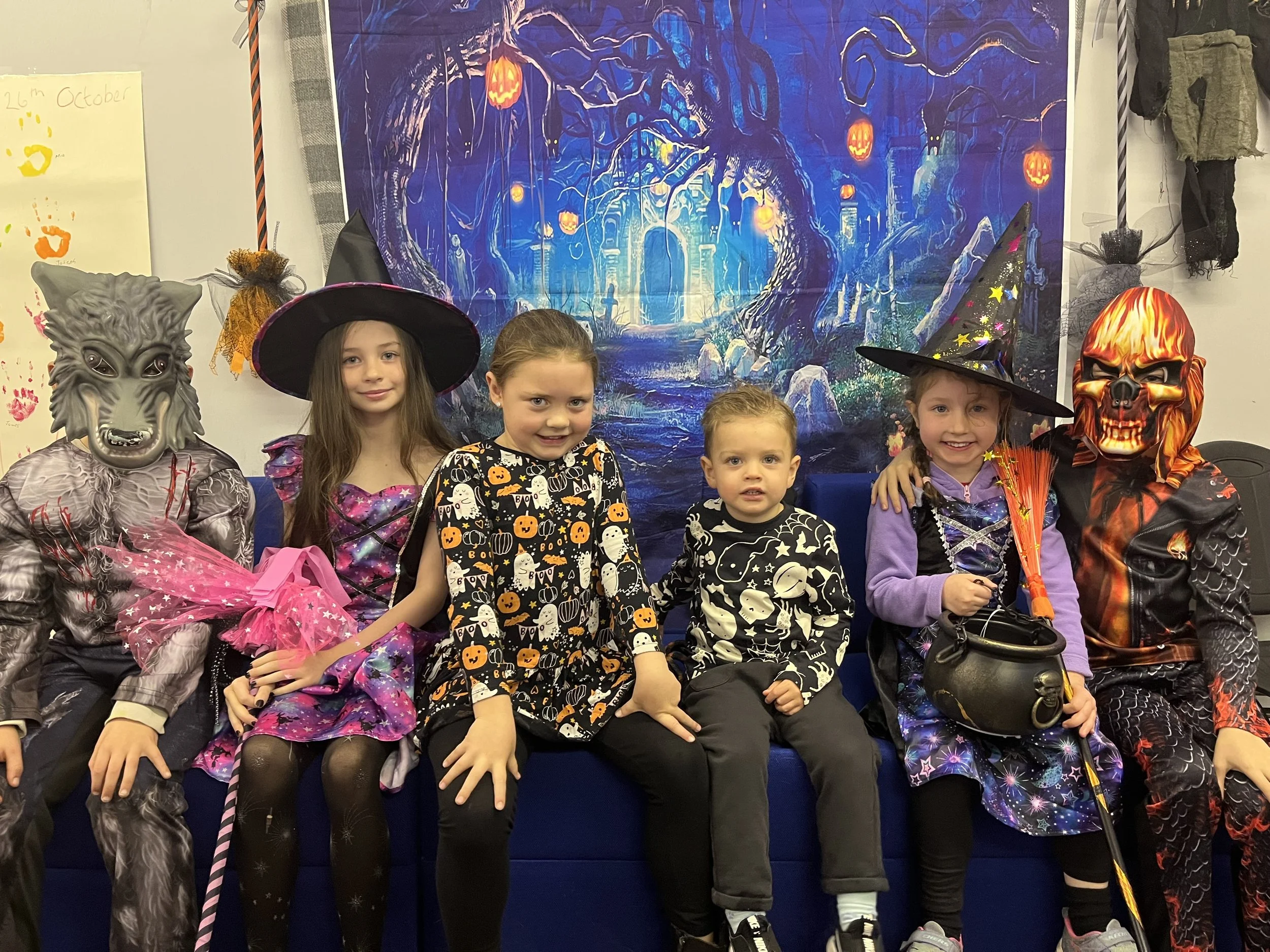 A group of five children dressed in Halloween costumes sitting together in front of a spooky forest backdrop with glowing jack-o'-lanterns, dressed as a wolf, a witch, and a skeleton, celebrating Halloween.