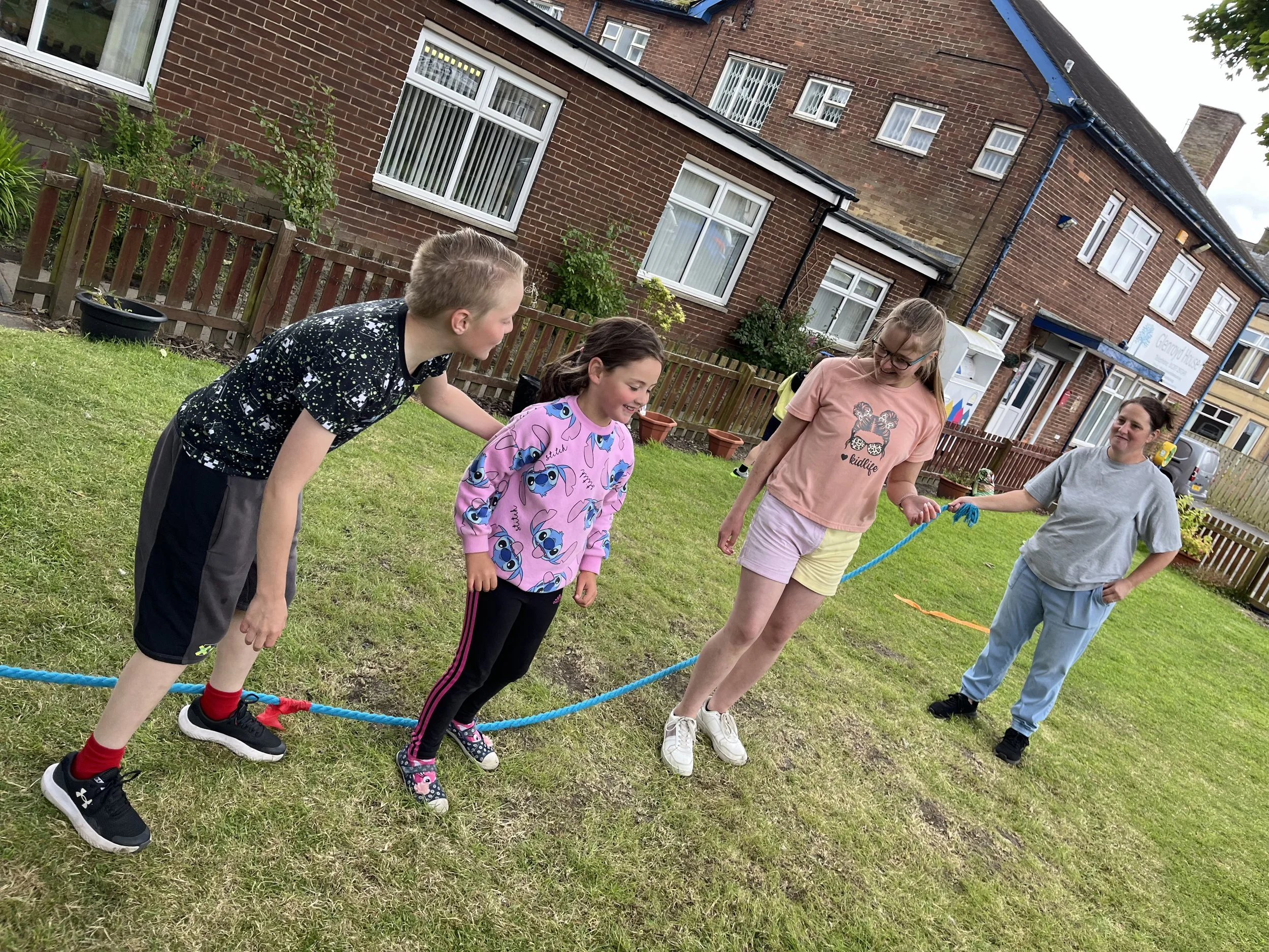A group of children and a woman outdoors, standing on a grassy area behind a blue rope, with brick residential buildings in the background. The children are smiling and appear to be enjoying an activity together.