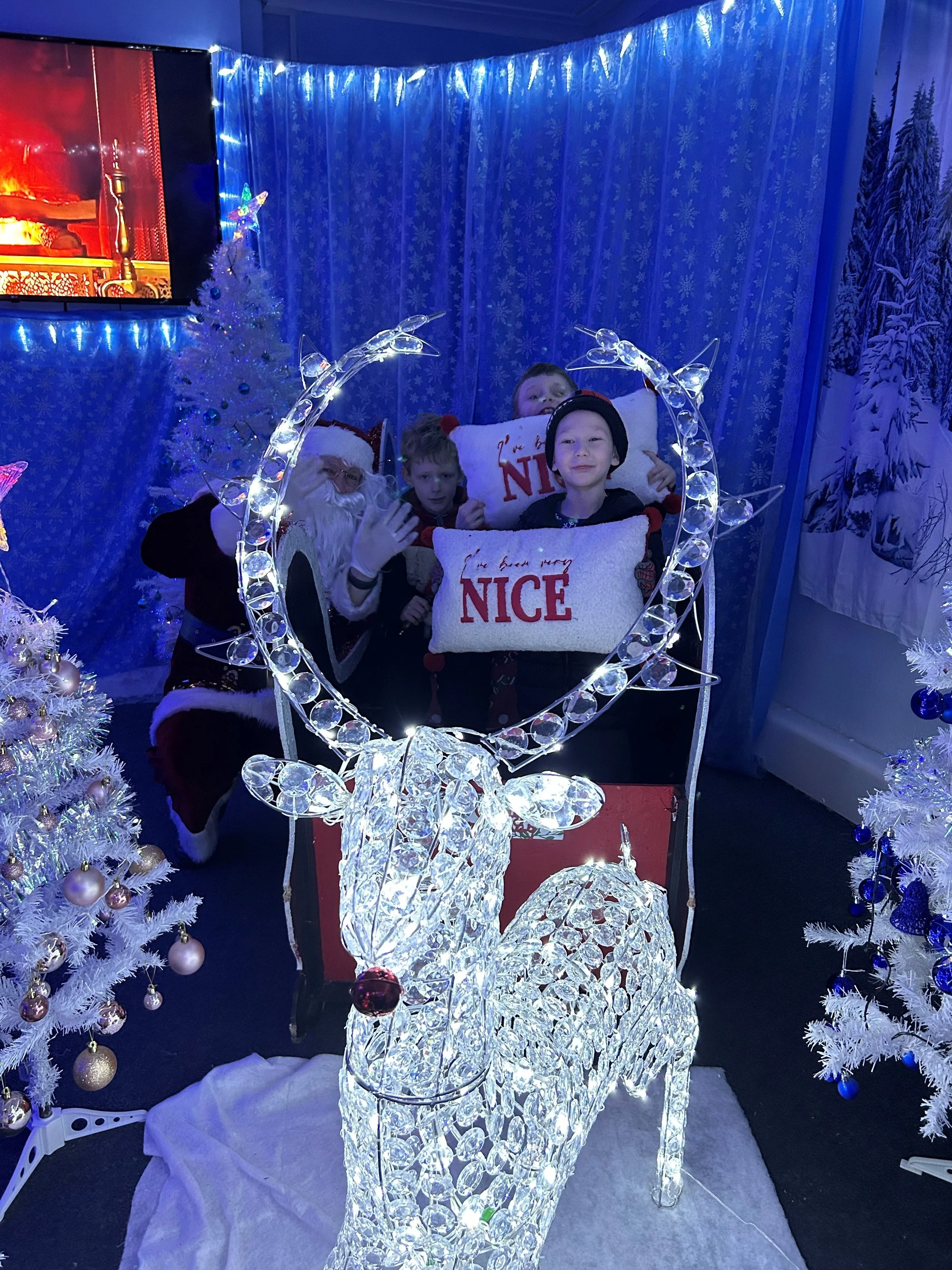 Children visiting Santa Claus with Christmas decorations, including white Christmas trees with ornaments, a lighted wire reindeer, and snowflake curtains.