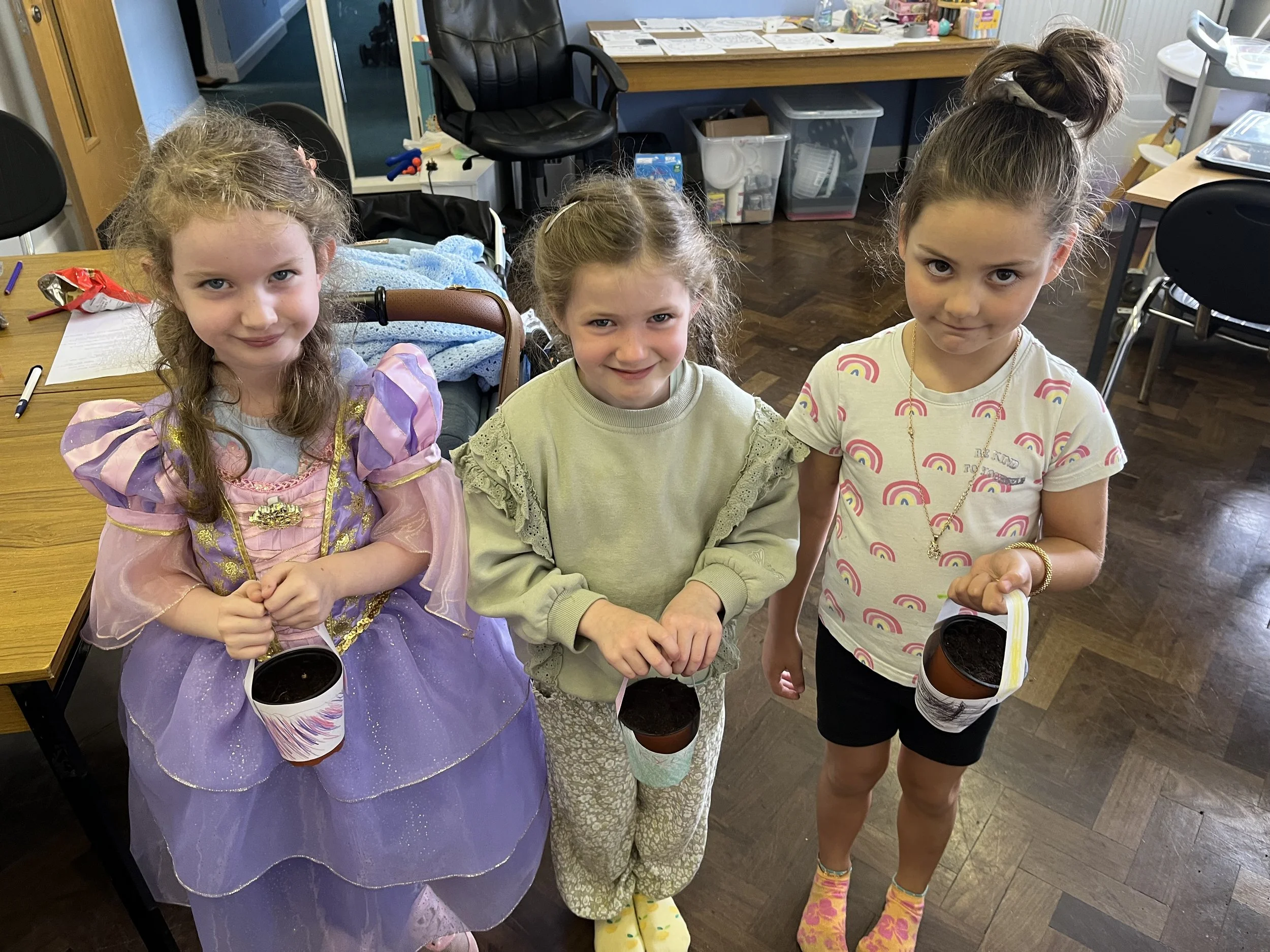 Three young girls standing together indoors holding cans of soda. One girl is dressed as a princess and another has pajamas with rainbows on them. They are smiling and looking at the camera.
