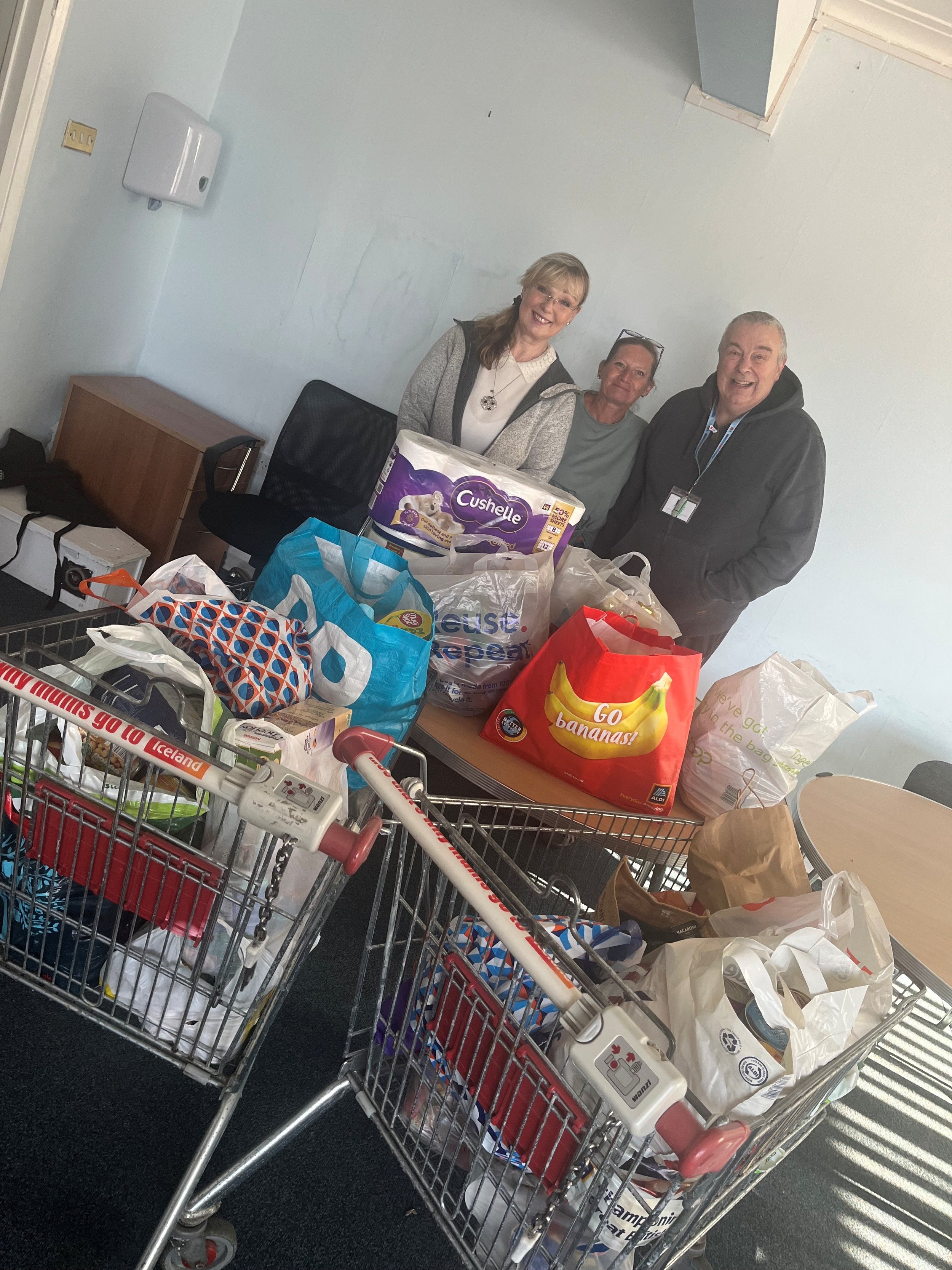 Three people standing behind a shopping cart filled with various grocery bags and items inside a store. They are smiling and looking at the camera.