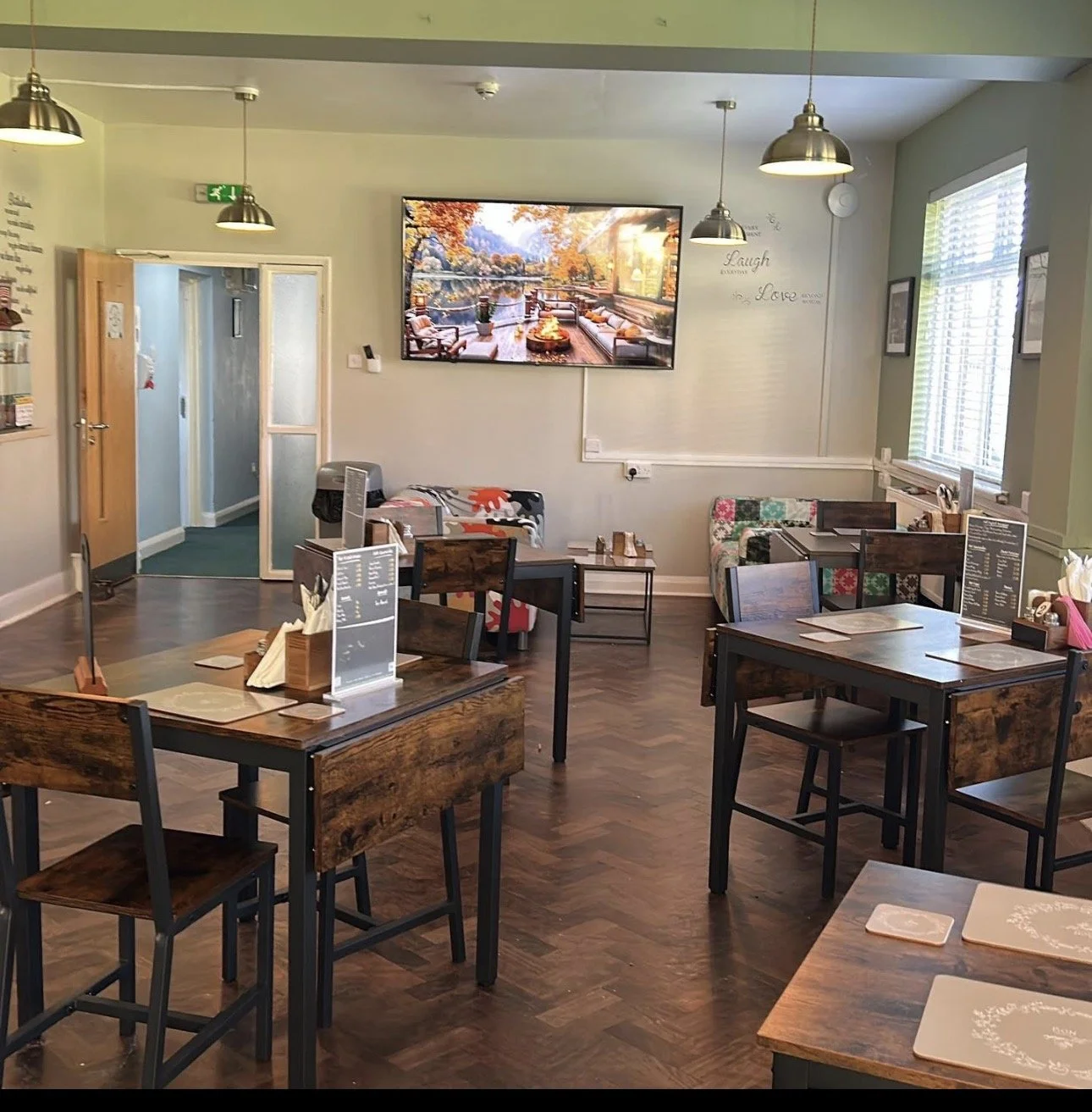 A cozy restaurant interior with wooden tables and chairs, a wall-mounted television displaying a nature scene, windows letting in natural light, and decorative wall words that say "Laugh" and "Love."