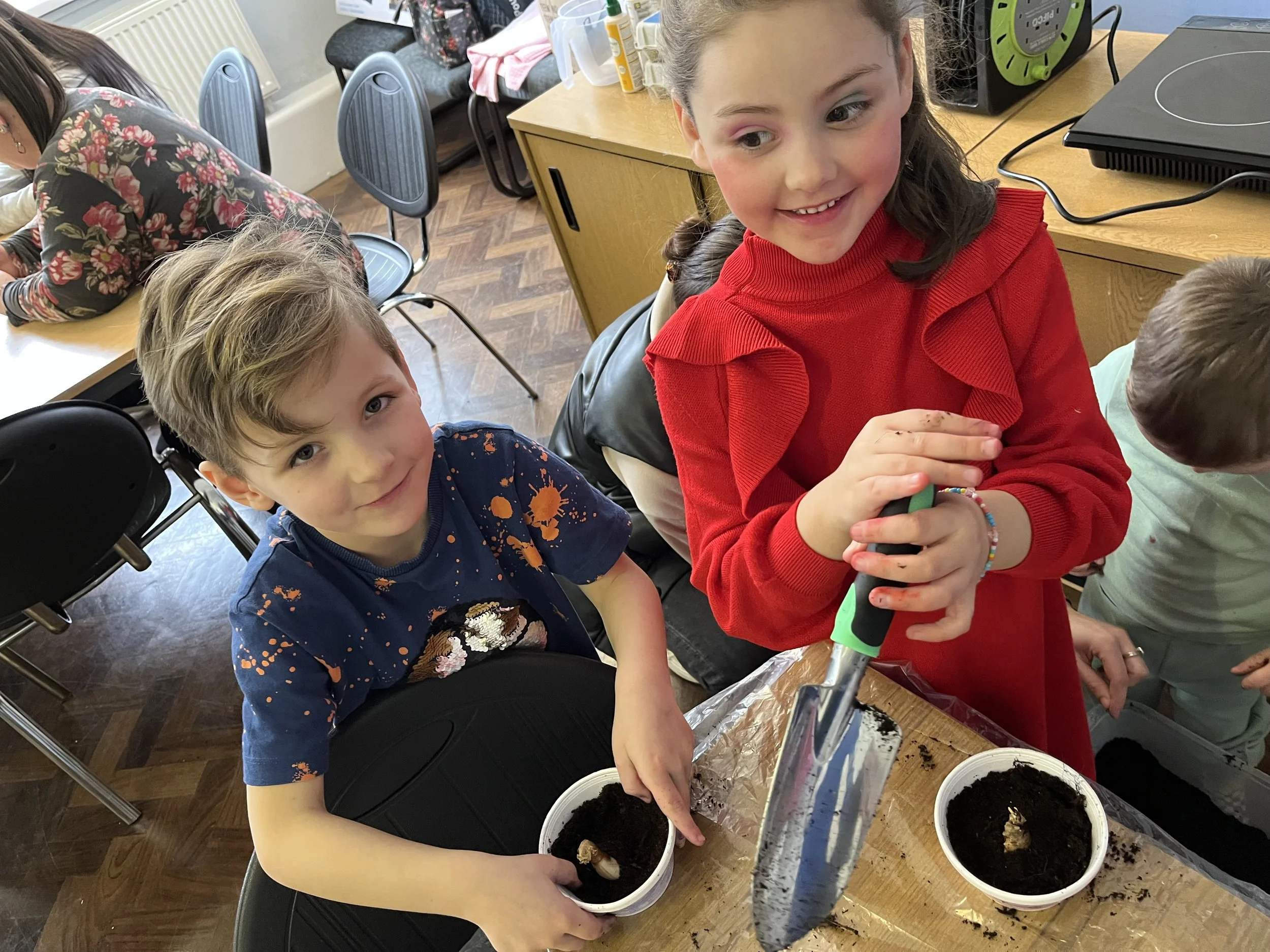 Children involved in a planting activity, filling small containers with soil using a scoop inside a classroom.