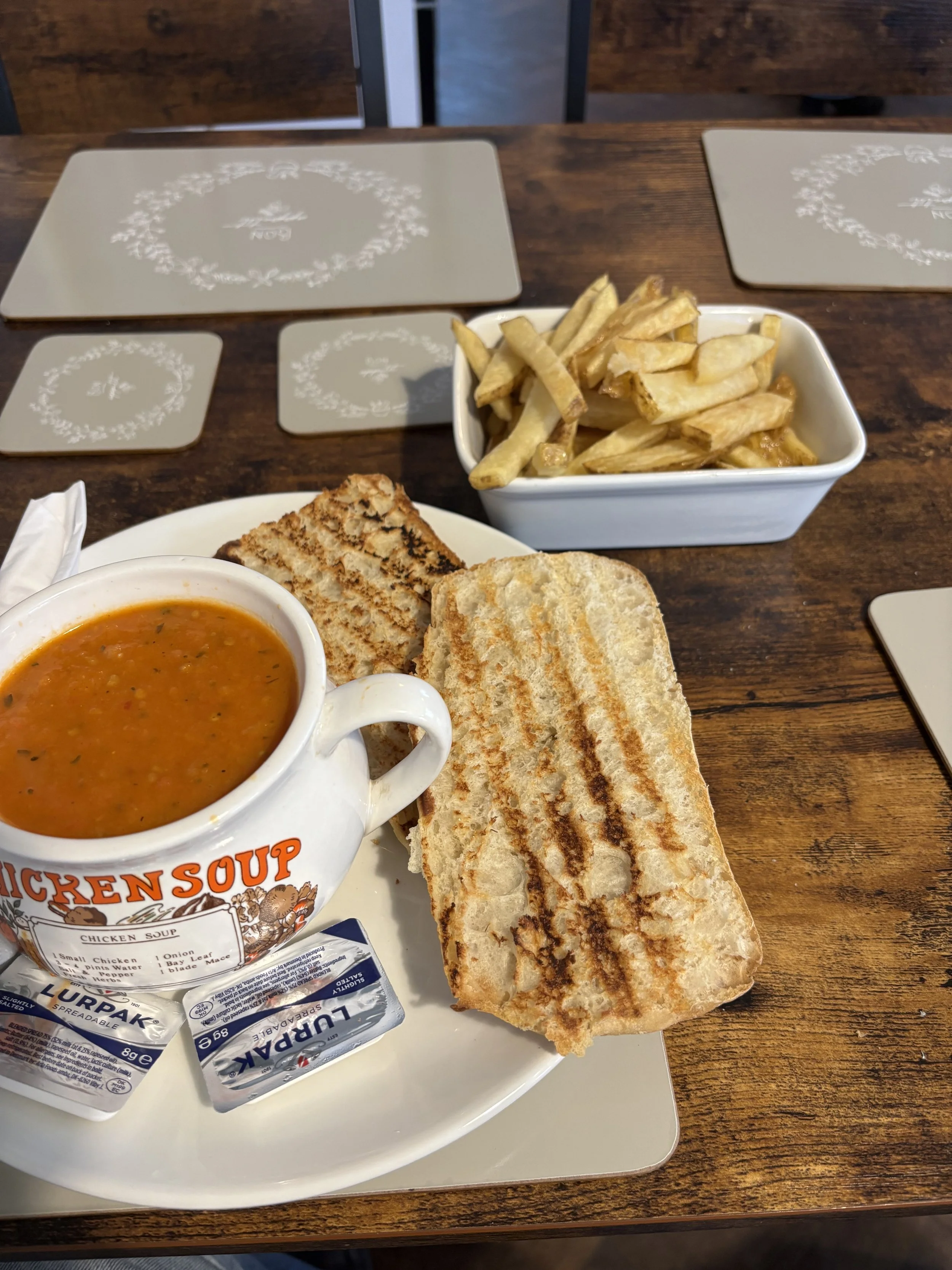 A meal consisting of a cup of chicken soup, two packets of bread, a slice of grilled cheese sandwich, French fries in a white bowl, and toasted bread on a white plate, on a wooden table.