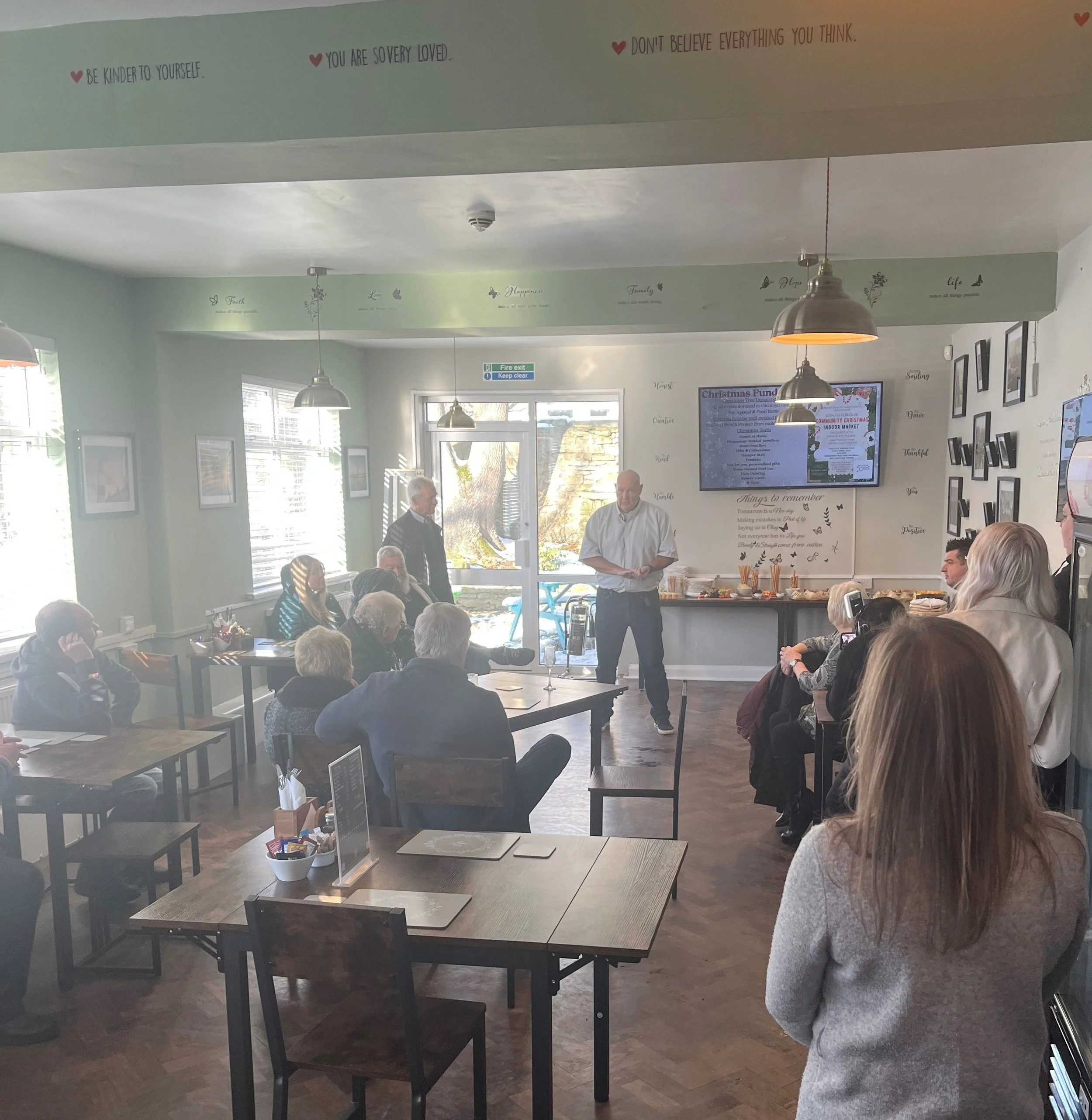 People gathering inside a room, sitting at tables, with a man standing in front, possibly giving a presentation or speech. The room has hanging lights, framed pictures on the wall, and a digital display showing a Christmas event.
