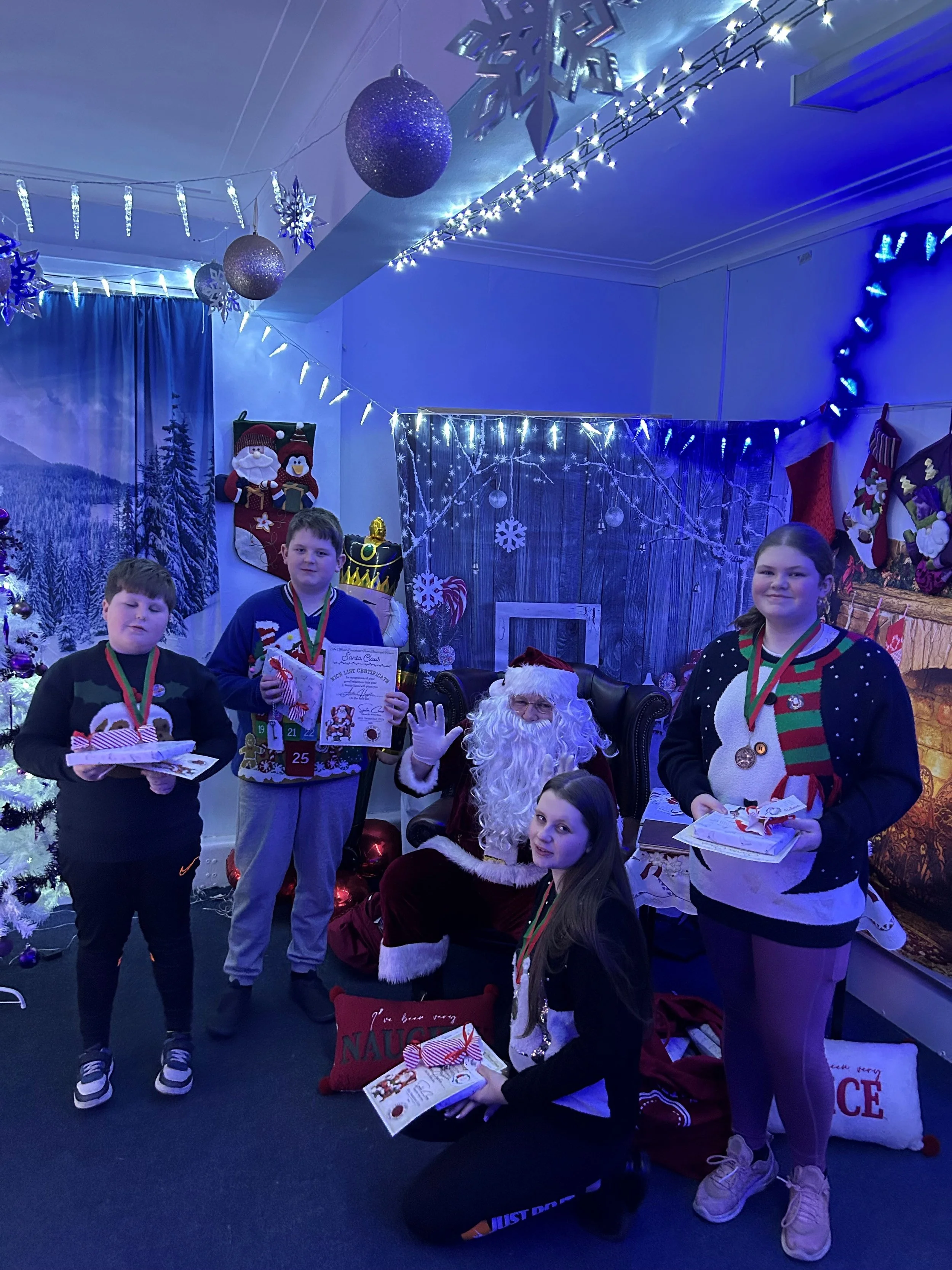 Children celebrating Christmas with an individual dressed as Santa Claus in a festively decorated room with Christmas stockings, lights, snowflakes, and holiday-themed decorations.