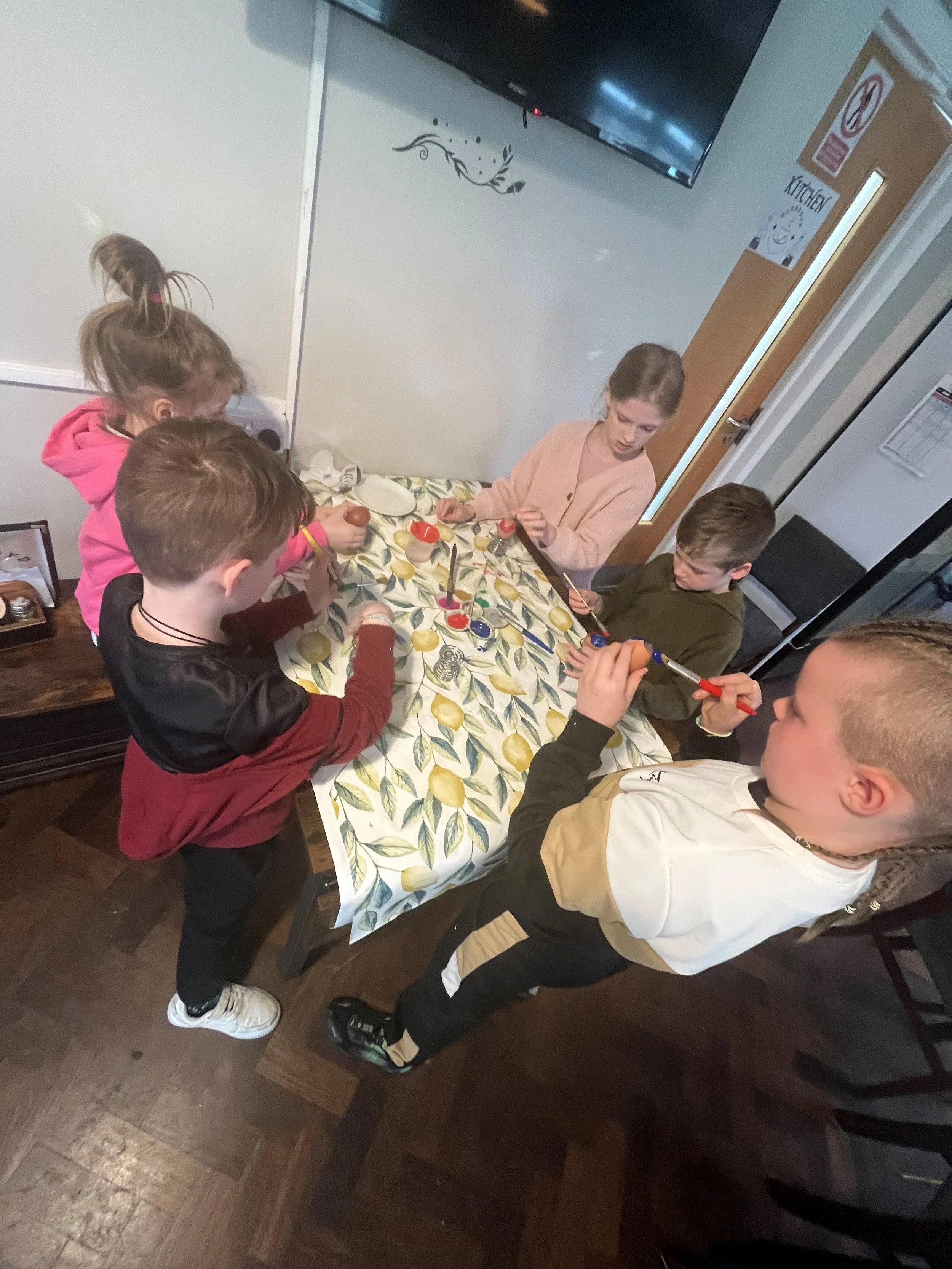 Five people, including two children and three adults, are gathered around a table painting Easter eggs with colorful dyes. The table has a lemon-patterned tablecloth.