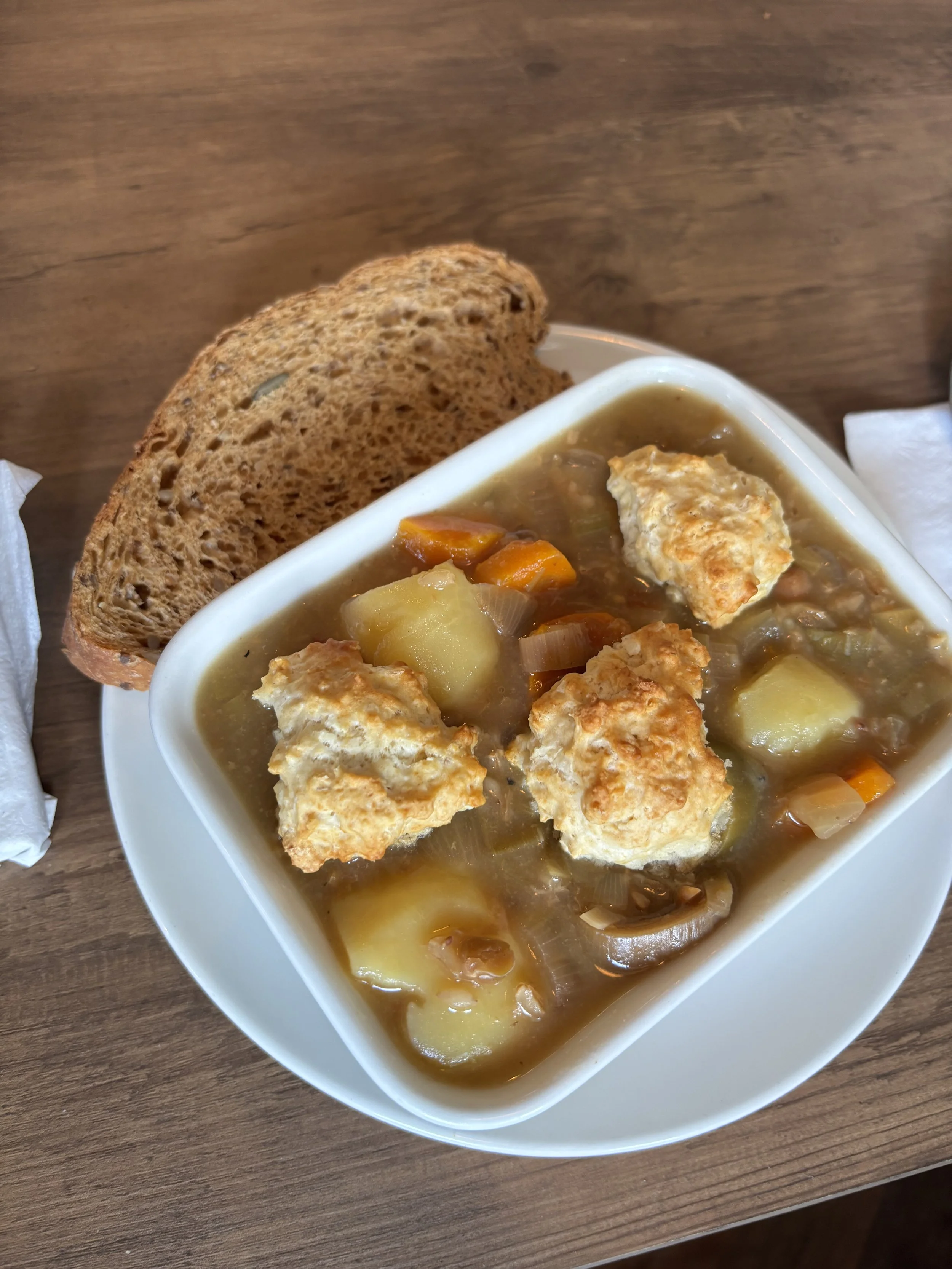 A bowl of chicken pot pie soup with chunks of chicken, potatoes, carrots, and celery, topped with pieces of baked bread, on a wooden table.
