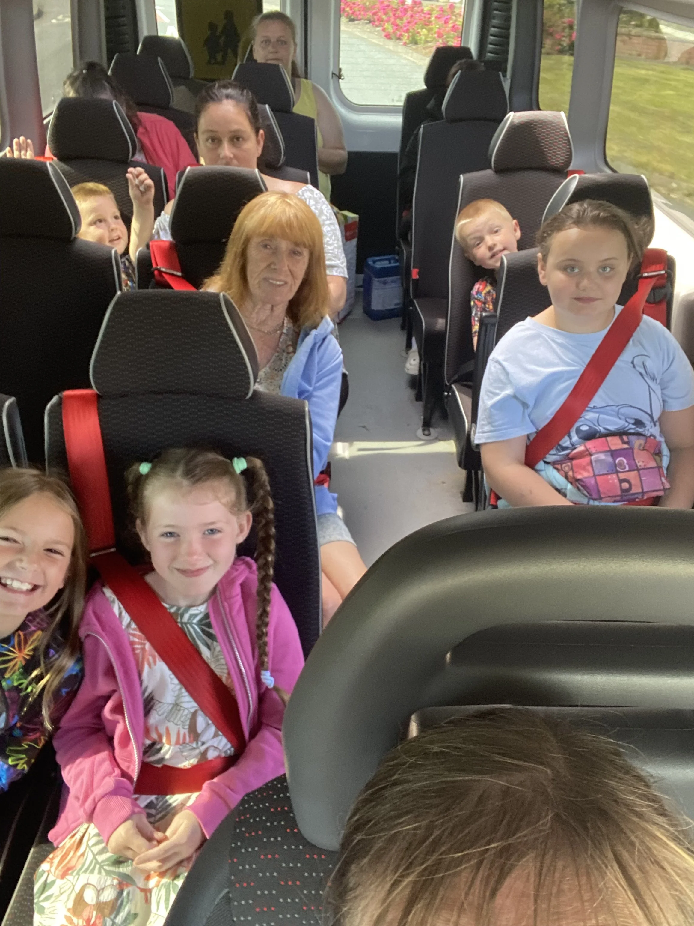 People seated in a bus with children and adults, some having their seat belts on, looking at the camera, with a window showing flowers outside.