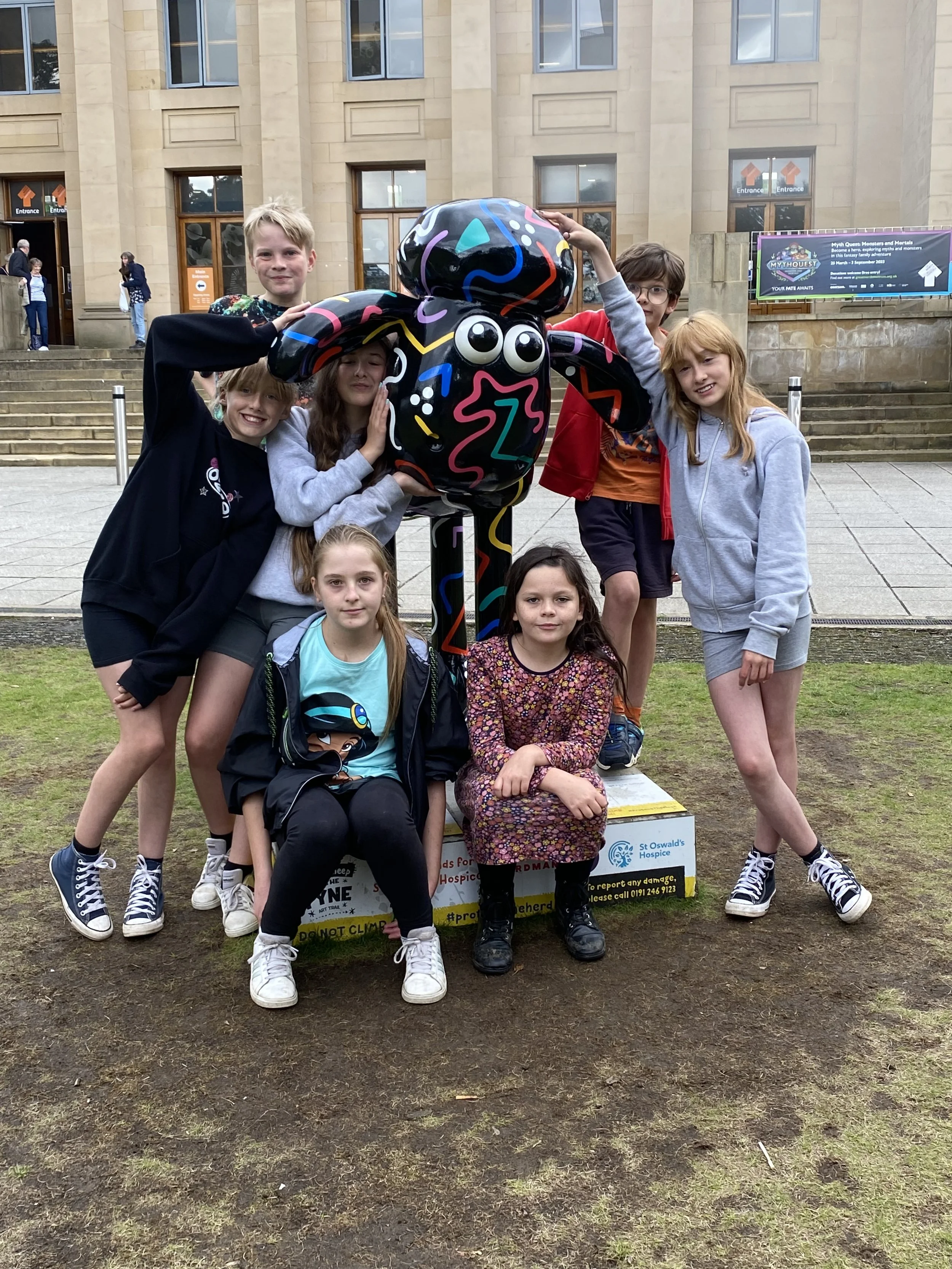 Group of seven children posing outdoors around a colorful black sculpture with large googly eyes, on a patch of muddy ground in front of a building with steps and a sign for St. Oswald's Hospice.