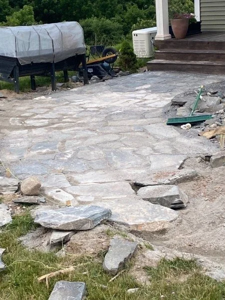 A partially finished stone pathway in a backyard with construction tools and materials nearby, next to a house with stairs and a potted plant.
