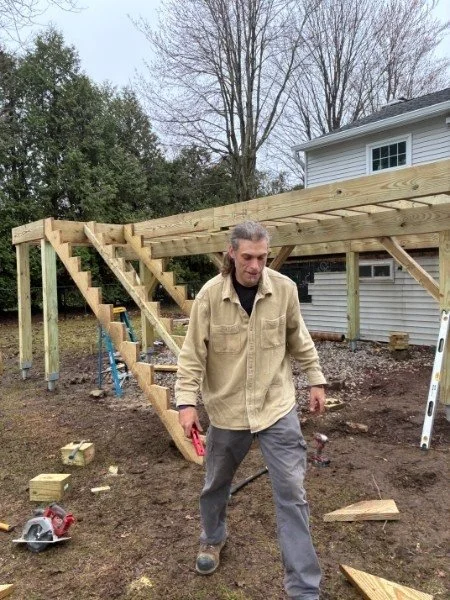 A man in a beige jacket and gray pants working on a construction site outdoors, with a partly built wooden deck or porch structure in the background, in a yard with trees and a white house.